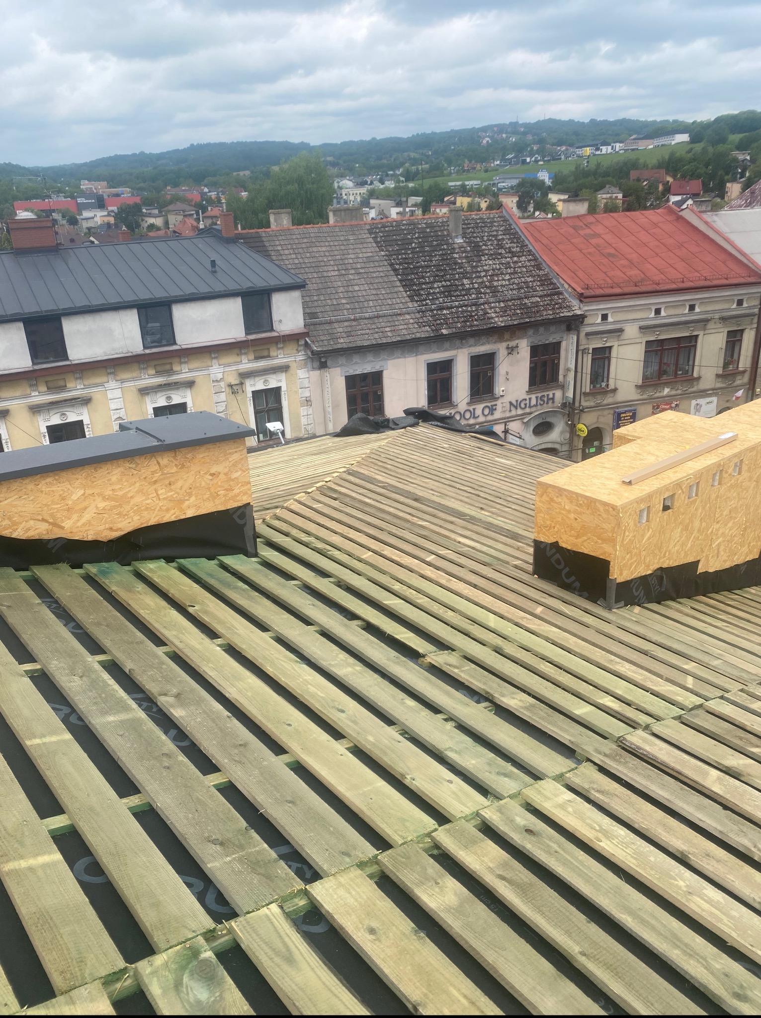 Konstrukcja dachu w trakcie budowy: drewniane łaty na czarnej membranie, w tle zabudowa miejska i pagórkowaty krajobraz pod pochmurnym niebem.