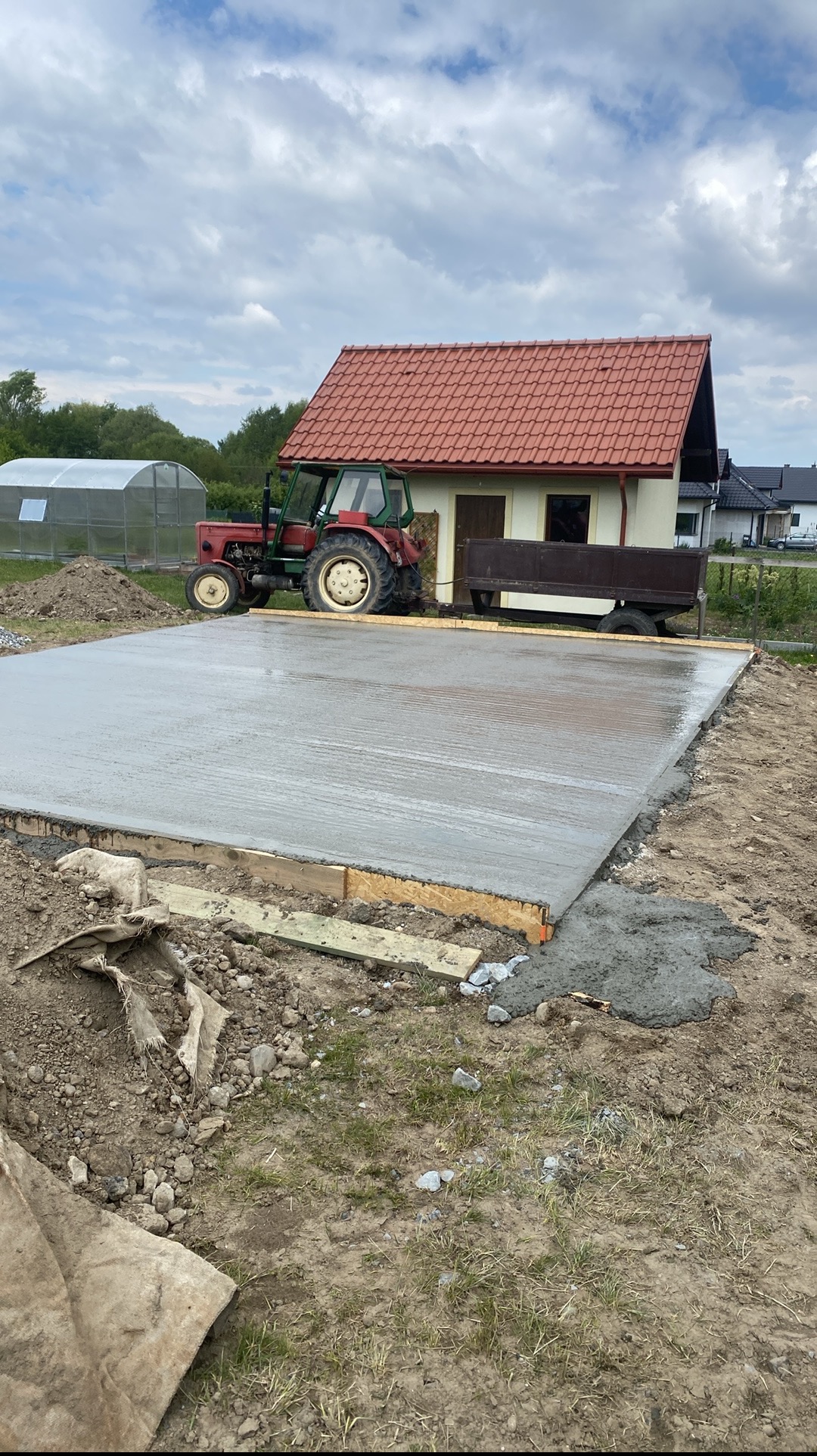 Wylana płyta fundamentowa na gruncie z szalunkiem drewnianym, w tle budynek z czerwonym dachem i traktor. Widok na świeży beton, dzień.