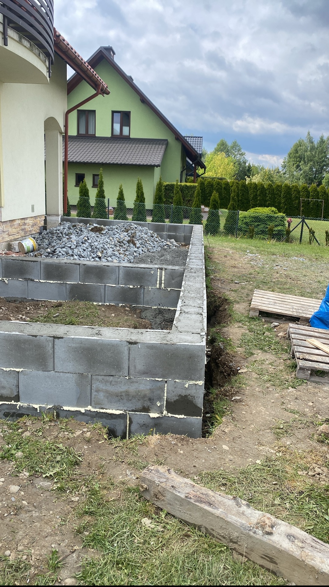 Budowa fundamentów z bloczków betonowych; widoczne szare bloczki, biała pianka montażowa, kamienie i zielony dom w tle.