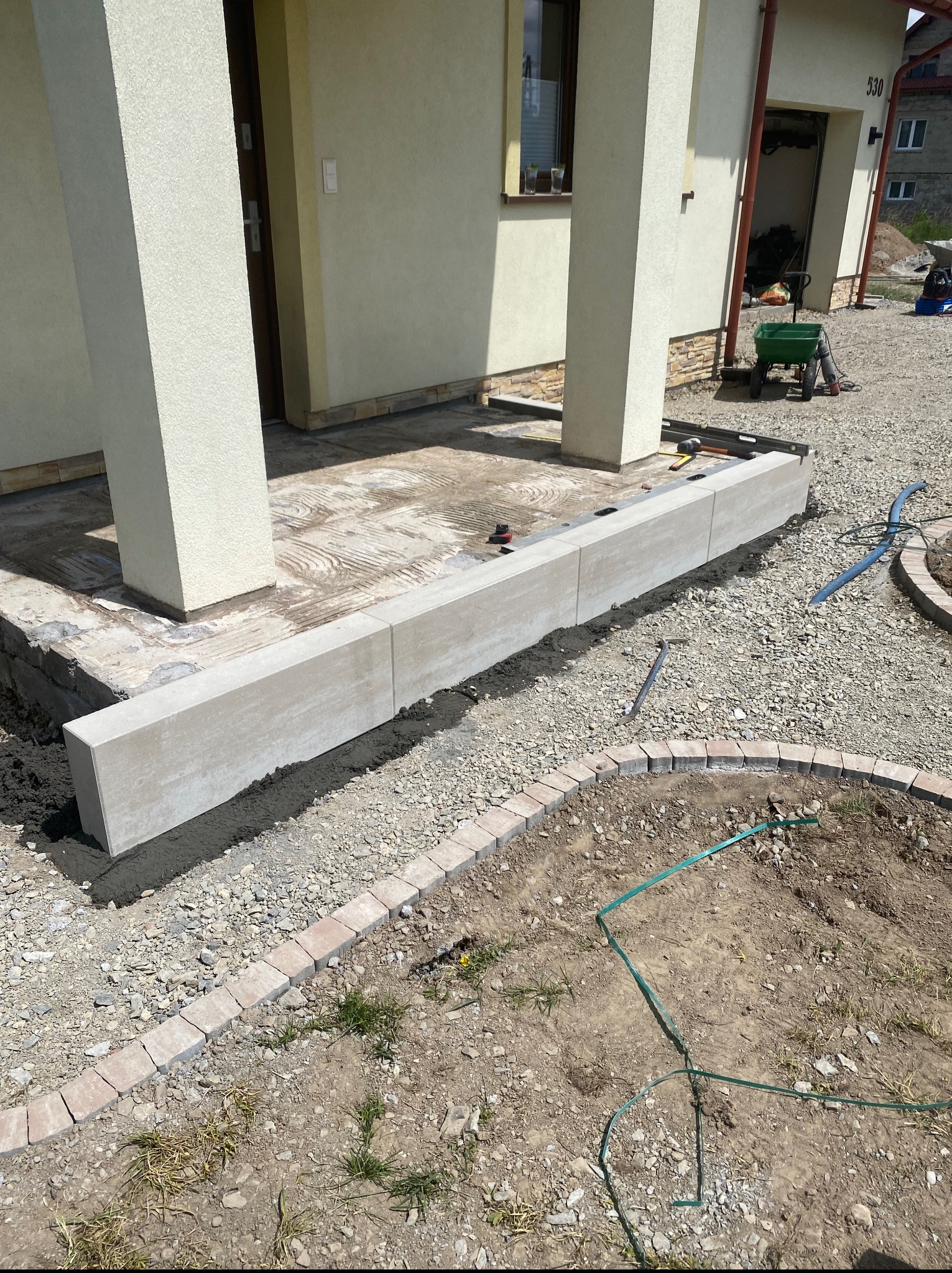 Montaż obrzeży betonowych przy tarasie domu jednorodzinnego. Widoczne narzędzia, piasek i kostka brukowa. Realizacja w trakcie prac wykończeniowych.