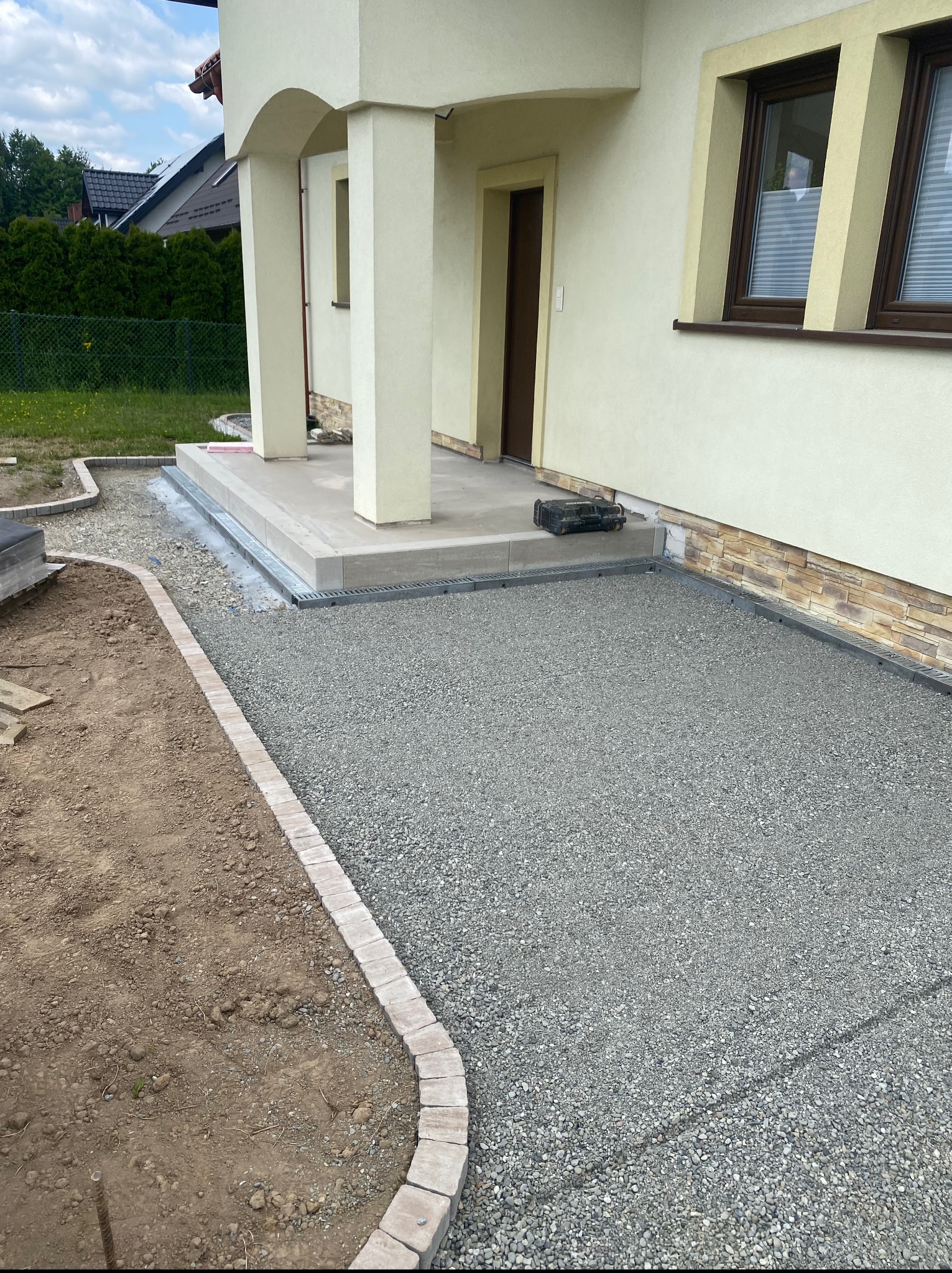 Wykończony podjazd z szarego żwiru, obrzeża z kostki. Widoczny fragment domu z kolumnami i oknem. Etap prac ziemnych przy układaniu nawierzchni.