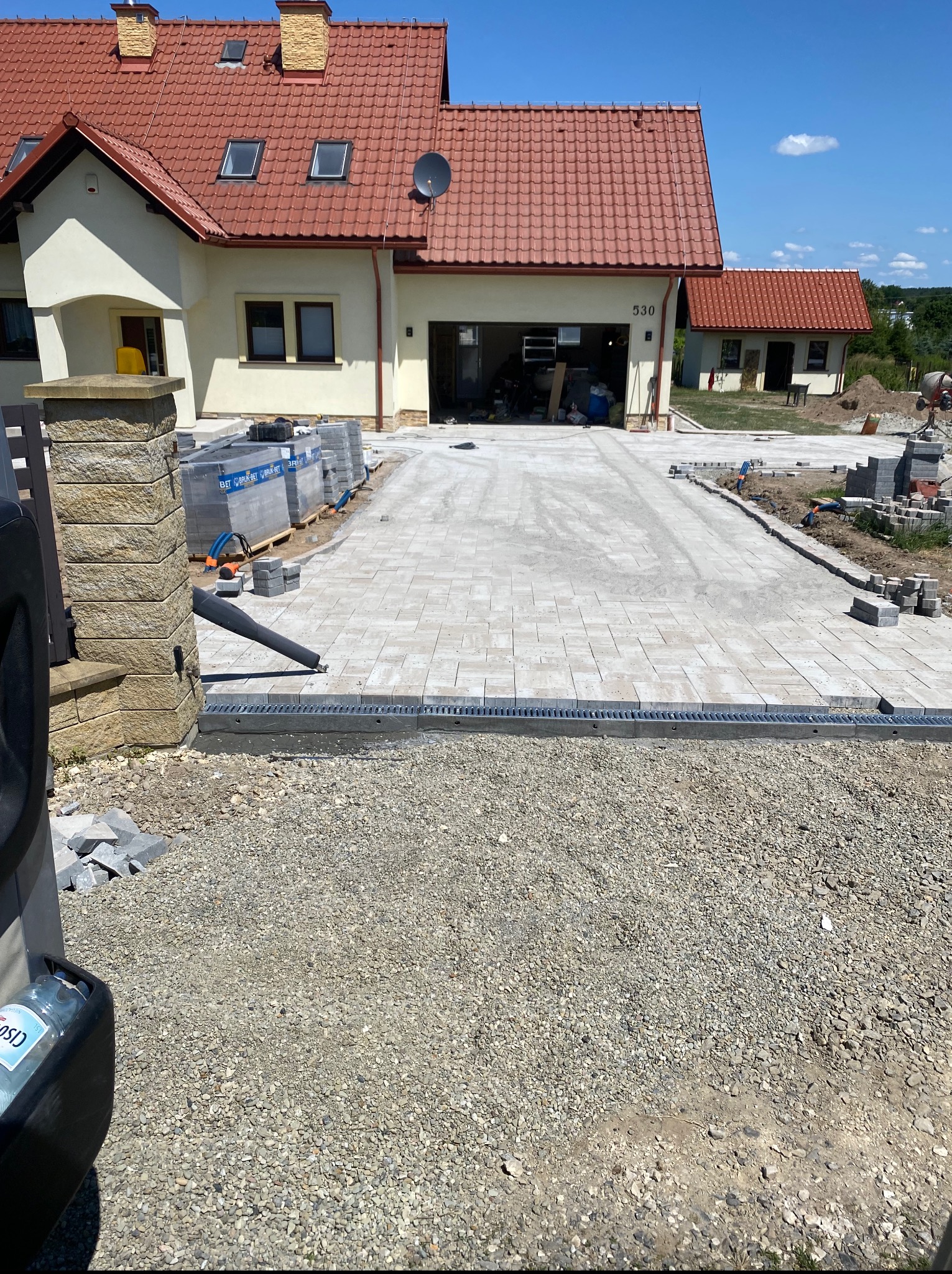 Układanie kostki brukowej na podjeździe do domu jednorodzinnego z widocznym systemem odprowadzania wody. Częściowo ułożona kostka, palety z kostką w tle.