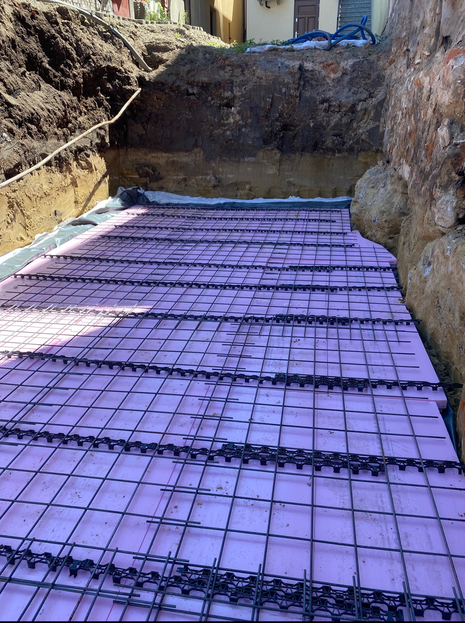 Przygotowanie podłoża pod płytę fundamentową: różowy styropian, siatka zbrojeniowa i czarne dystanse w wykopie. Widoczne ściany wykopu i biała folia izolacyjna.