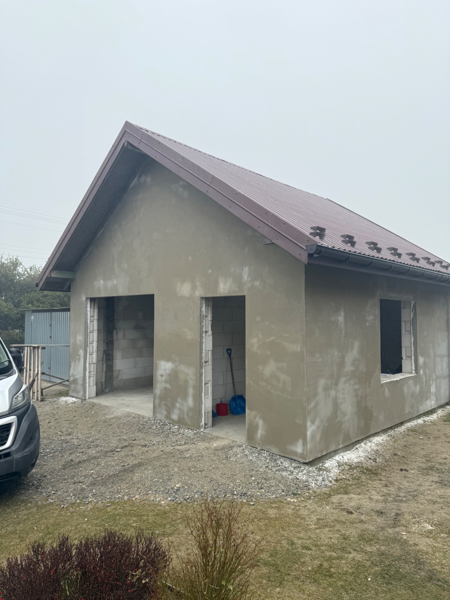 Surowy, otynkowany budynek gospodarczy z brązowym dachem, widoczne bloczki betonowe w otworach drzwiowych i okiennym, szpadel i wiadro wewnątrz. Szary dzień.