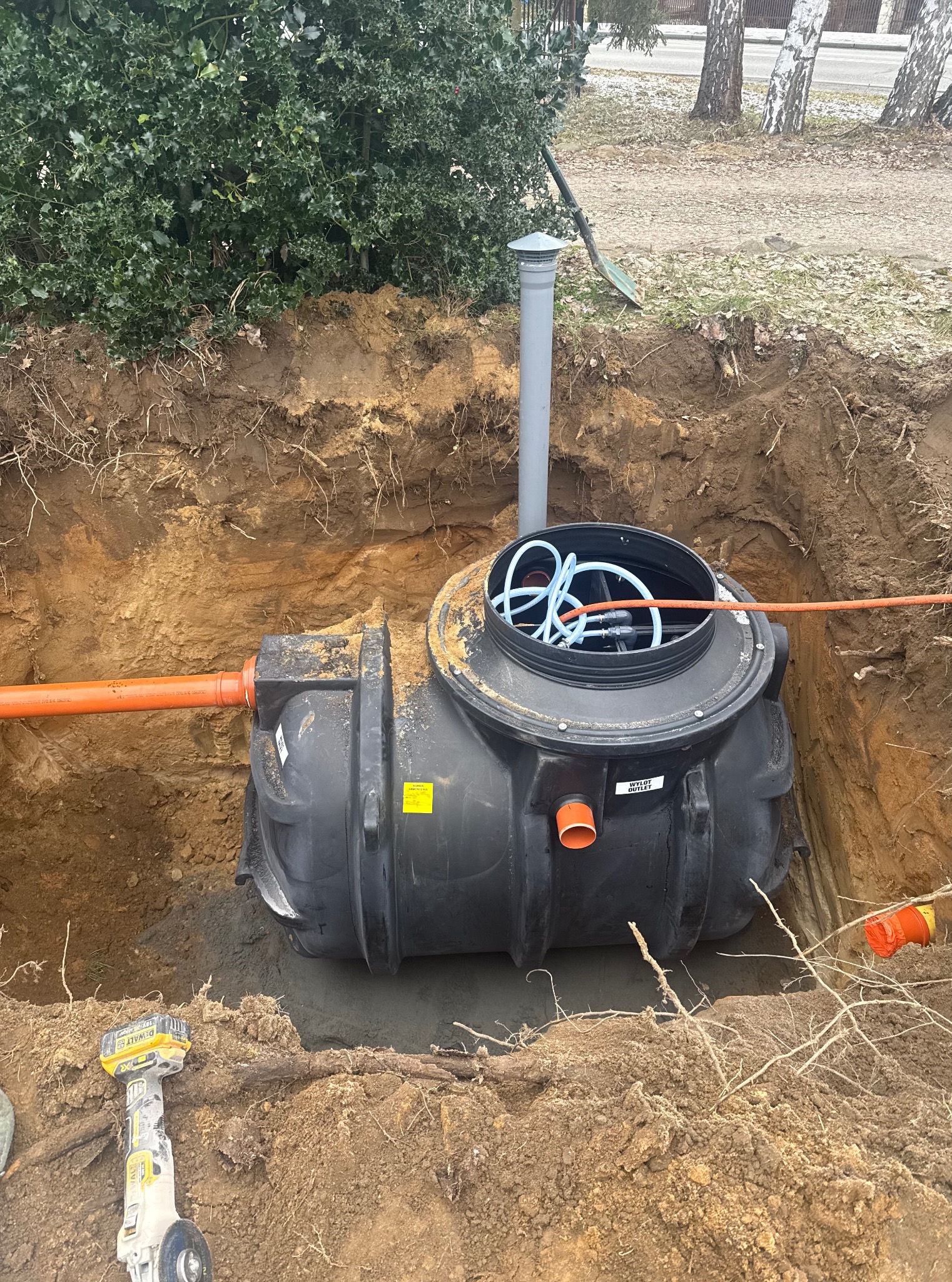 Czarna, dwuczęściowa przydomowa oczyszczalnia ścieków w wykopie, z widocznymi rurami doprowadzającymi i odprowadzającymi. W tle drzewa i krzewy.