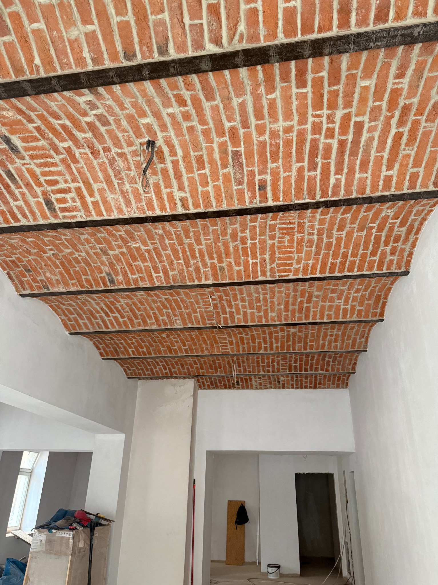 Sufit łukowy z czerwonej cegły, wzmocniony metalowymi belkami, w surowym wnętrzu. Widoczne przewody elektryczne i przygotowane ściany do malowania.