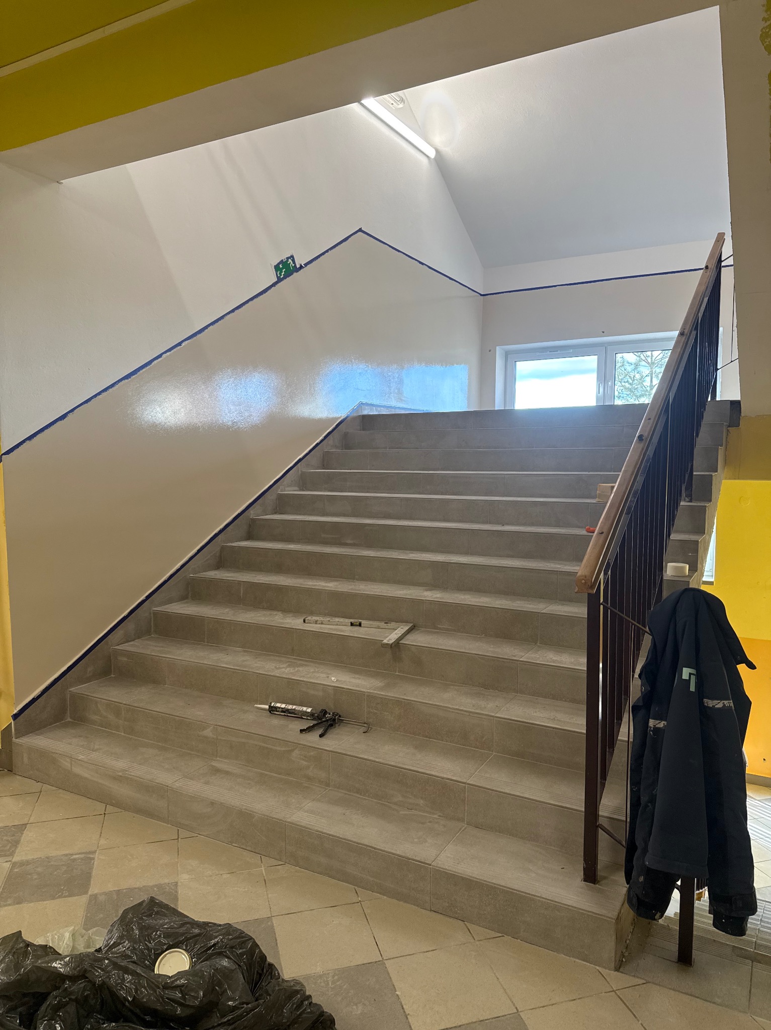 Klatka schodowa w trakcie malowania, zabezpieczona taśmą malarską. Widoczne narzędzia malarskie i częściowo pomalowana ściana. Na dole worki na śmieci.
