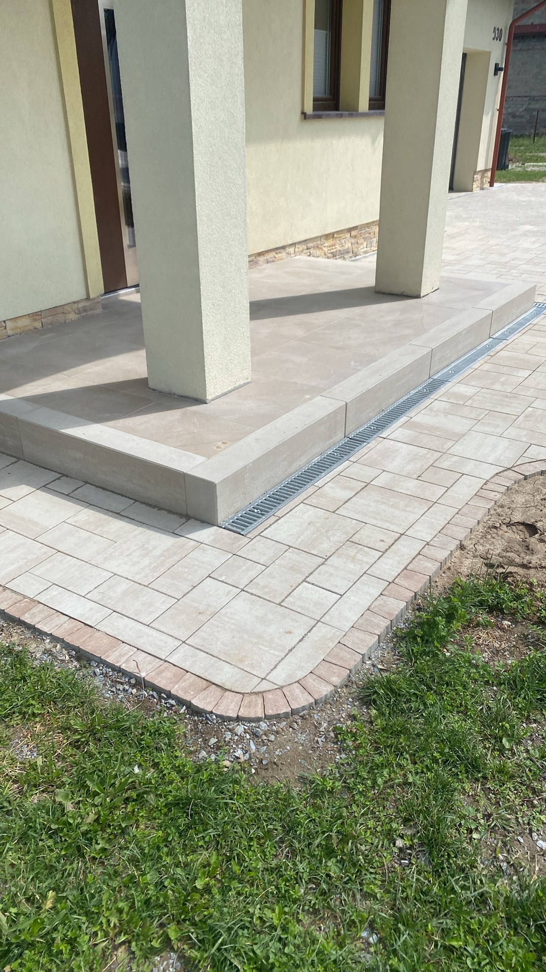 Wykończony taras z kostki brukowej w odcieniach beżu i szarości, z odpływem liniowym, prowadzący do wejścia domu z kolumnami. Widok z poziomu gruntu.