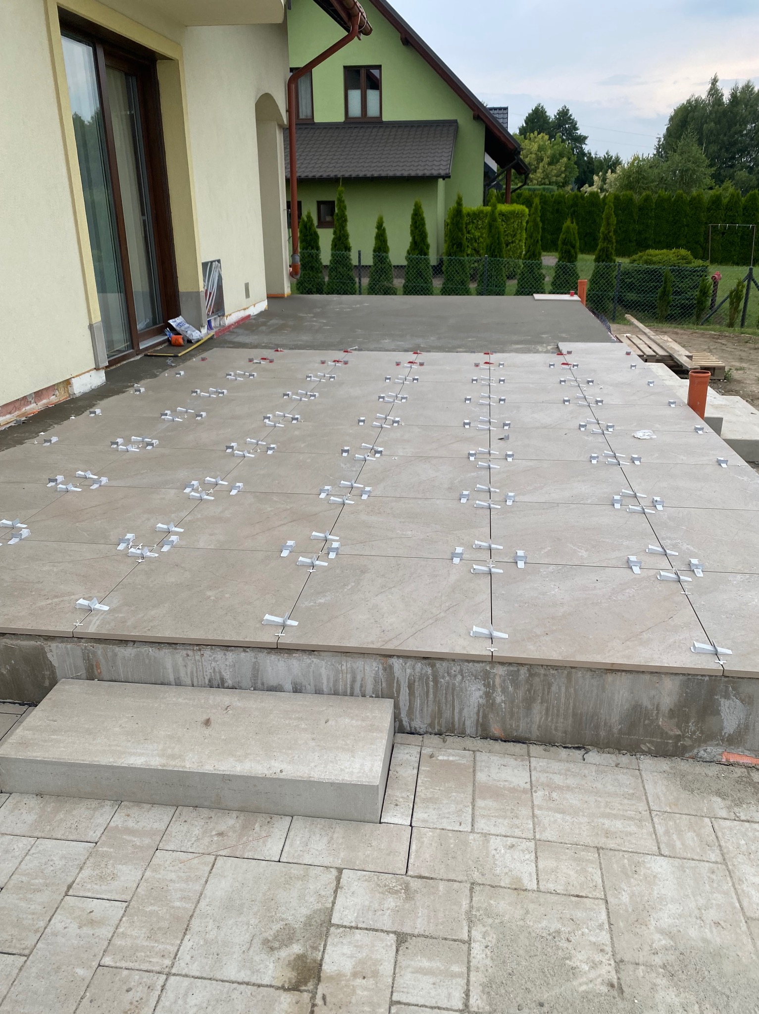 Układanie płytek na tarasie z użyciem klipsów poziomujących. Widoczne płytki w odcieniach beżu, fragment domu i ogrodu w tle. Prace wykończeniowe na zewnątrz budynku.