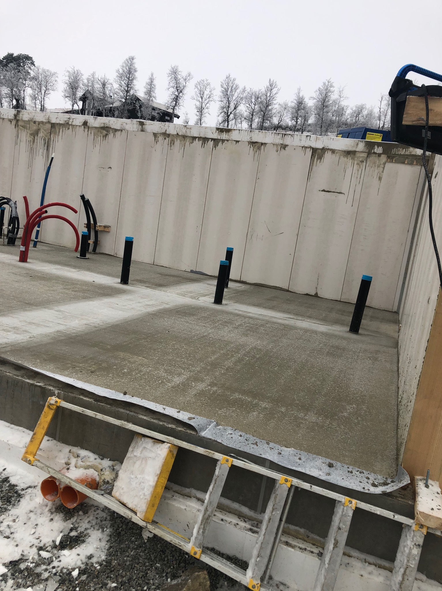 Zalany fundament budynku żelbetowego z wystającymi rurami instalacyjnymi. W tle ściany szalunkowe i zimowy krajobraz. Na pierwszym planie drabina.
