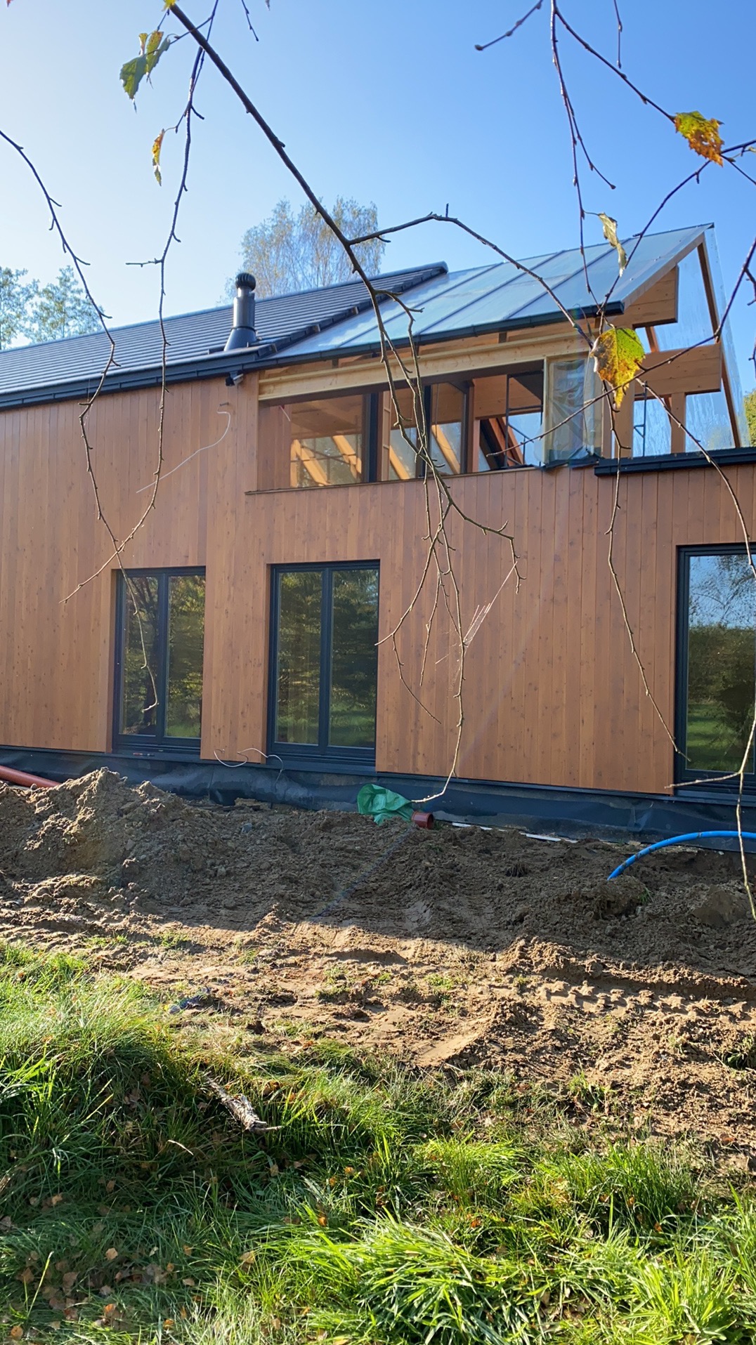 Nowoczesny dom z drewnianą elewacją i dużymi oknami, częściowo zasłonięty gałęziami drzew. Widoczne prace ziemne wokół budynku i instalacje.