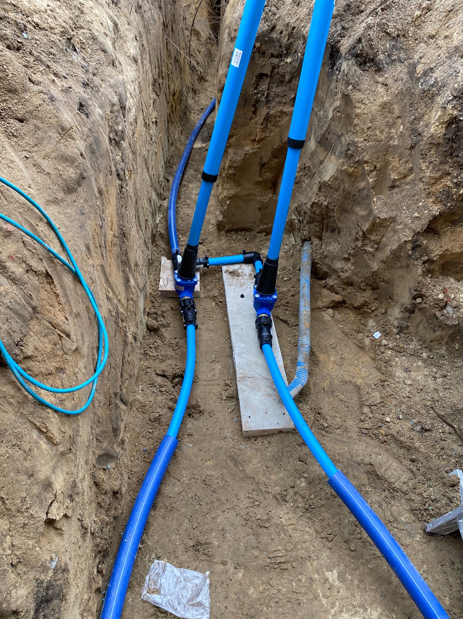Niebieskie rury instalacji wodnej w wykopie ziemnym, połączone złączkami na drewnianych podkładkach. Widoczne ściany wykopu i fragment drabiny.