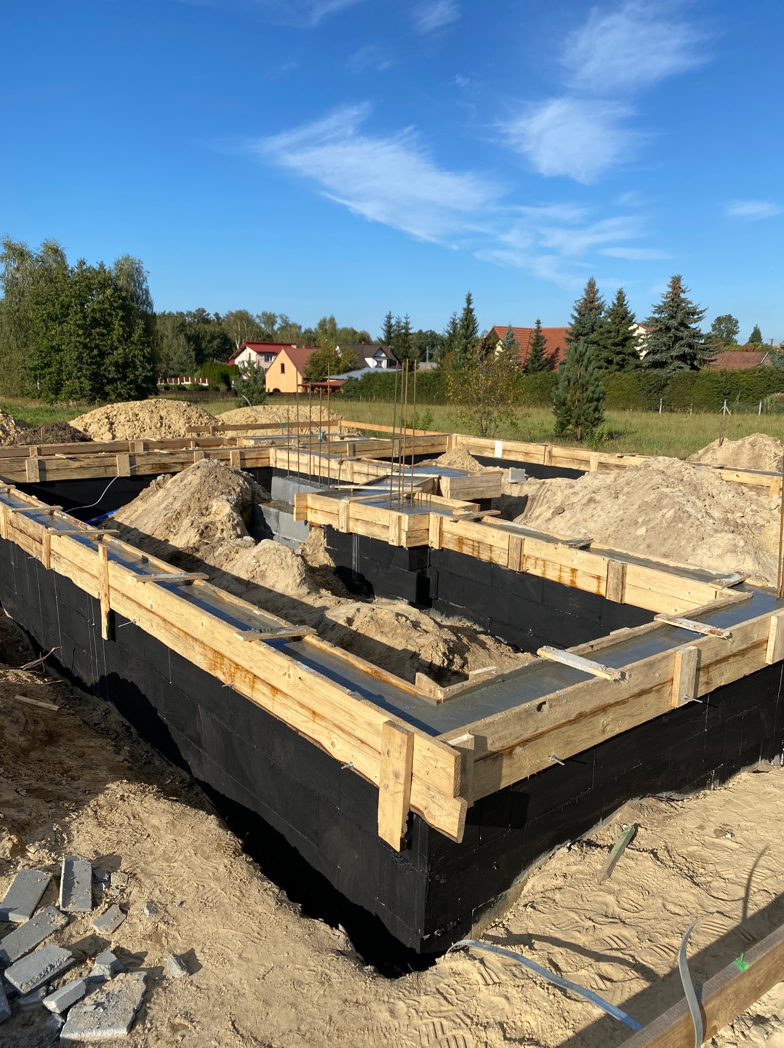 Zalane betonem szalunki fundamentów domu jednorodzinnego, zabezpieczone czarną folią. Widoczne zbrojenia i nasypany piasek. W tle zabudowa jednorodzinna i błękitne niebo.