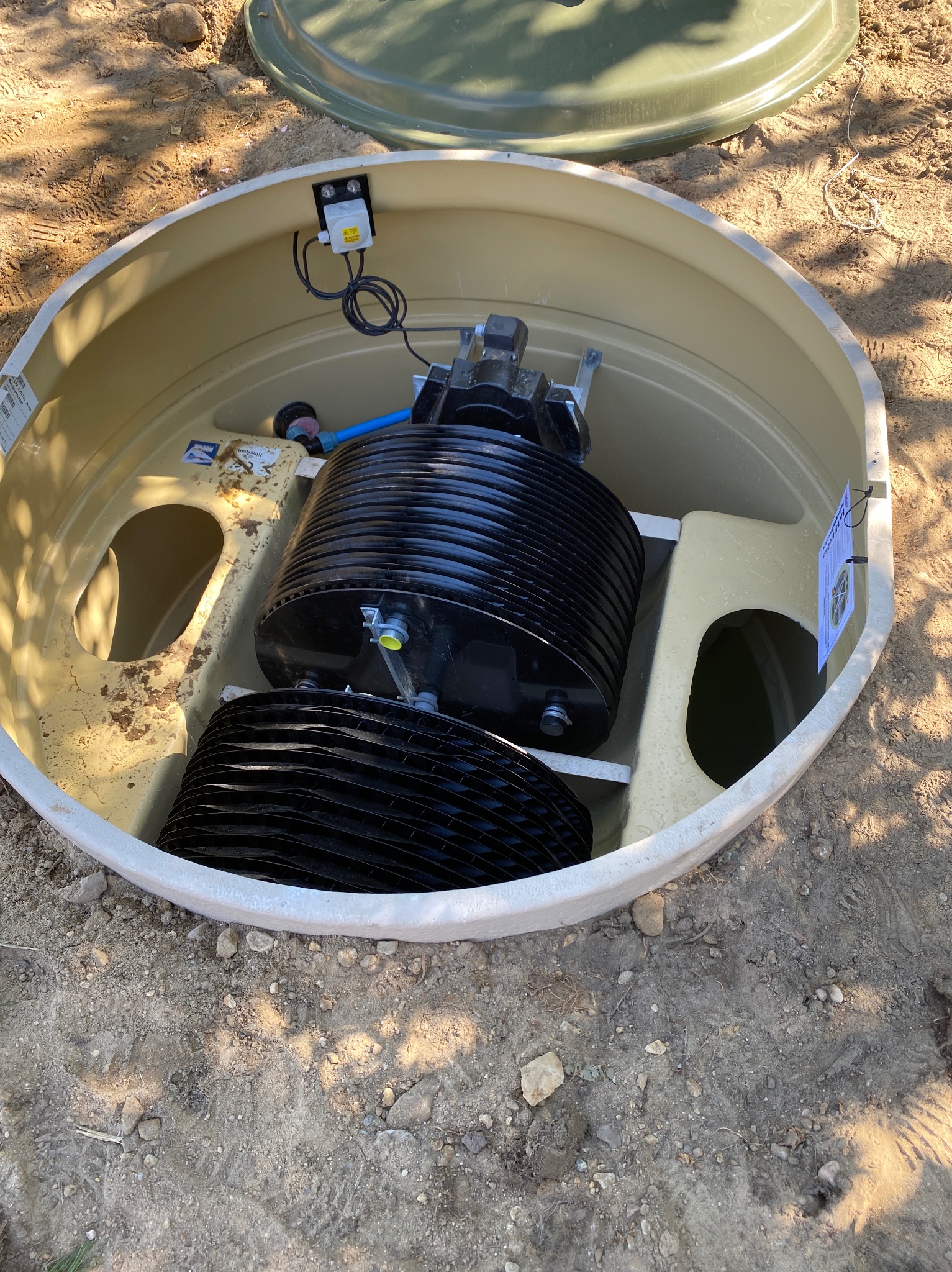 Wnętrze beżowego zbiornika oczyszczalni ścieków z czarnymi elementami filtracyjnymi, widoczne przewody i pokrywa obok. Ziemiste tło.