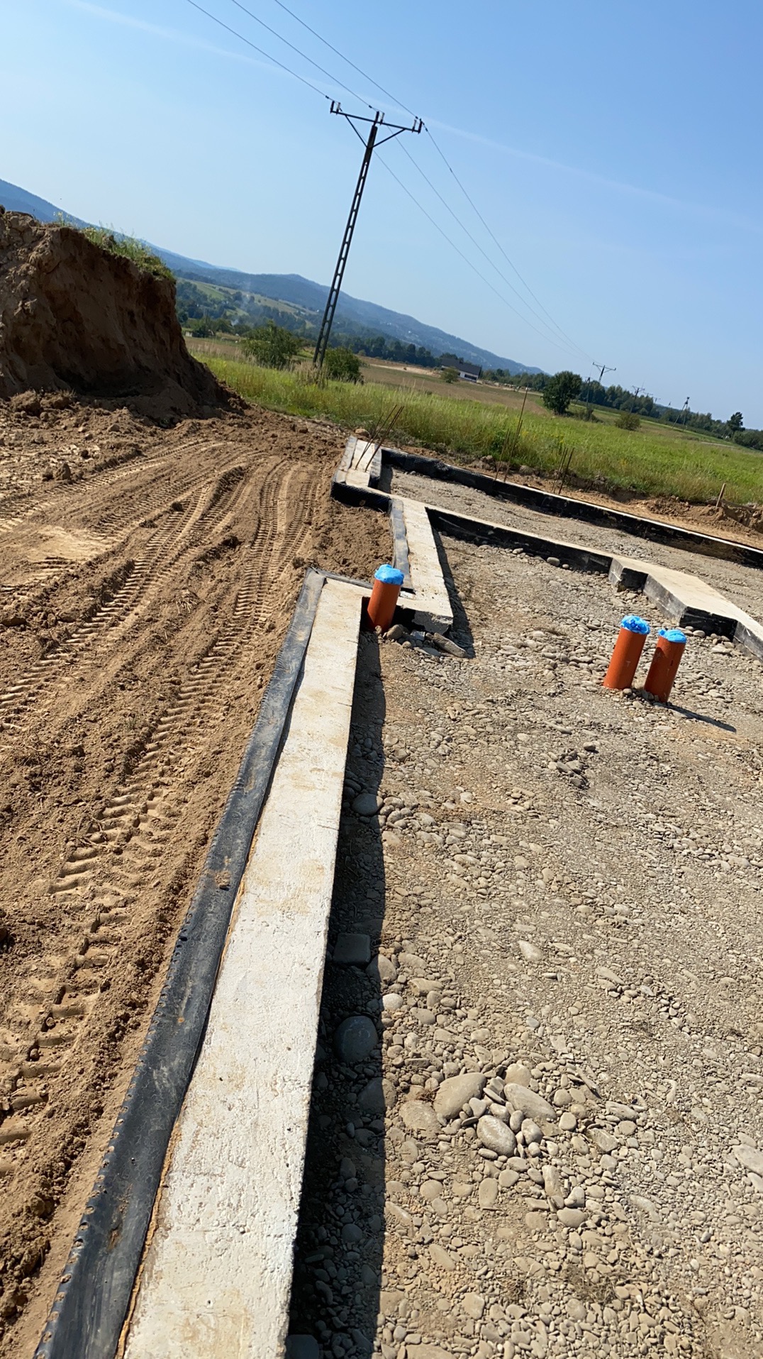 Fundamenty domu jednorodzinnego w Mokrzysku, z widocznymi elementami izolacji i pionowymi rurami kanalizacyjnymi. W tle pagórkowaty krajobraz.