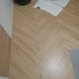 Ułożone panele podłogowe w jodełkę, widok z góry, z koszem na śmieci i białą szmatką. Jasne, ciepłe odcienie drewna.