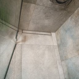 Krystian Kis - Wykończona kabina prysznicowa z odpływem liniowym i szklaną ścianą. Płytki w odcieniach szarości, na ścianie dekoracyjny wzór. Metalowy koszyk na akcesoria.