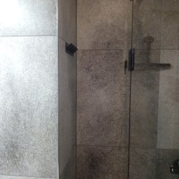 Krystian Kis - Wykończona kabina prysznicowa z szarymi, matowymi płytkami. Widoczne mocowania szklanych drzwi i szczotka do czyszczenia na ścianie. Minimalistyczny design.