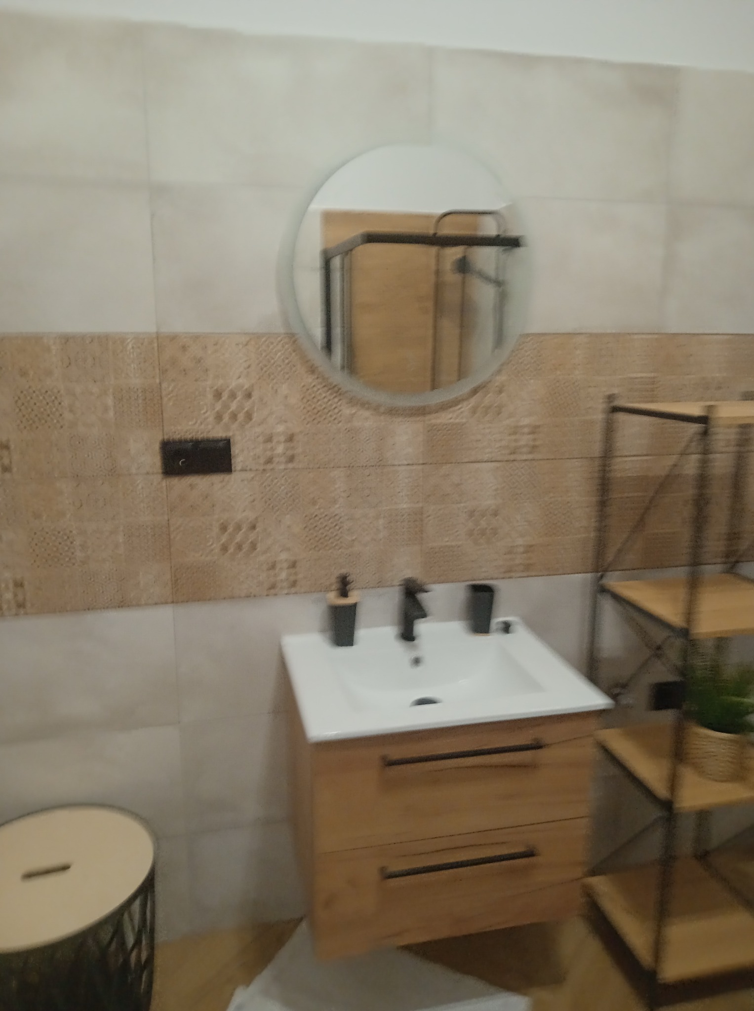 Wykończona łazienka: biała umywalka z drewnianą szafką, okrągłe lustro. Na ścianie beżowe płytki z wzorem i włącznik. W tle regał z drewnianymi półkami.