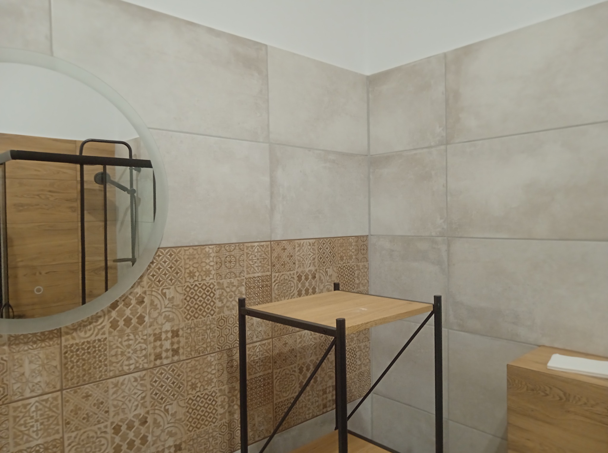 Wykończona łazienka z beżowymi płytkami: gładkie na górze, wzorzyste na dole. Okrągłe lustro odbija kabinę prysznicową. Drewniana szafka i metalowy regał.