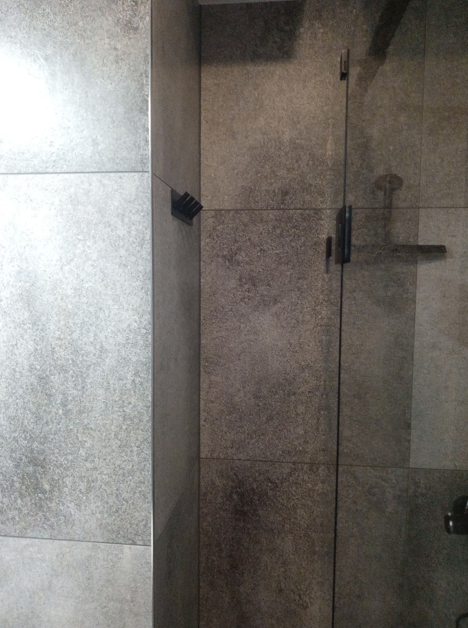 Wykończona kabina prysznicowa z szarymi, matowymi płytkami. Widoczne mocowania szklanych drzwi i szczotka do czyszczenia na ścianie. Minimalistyczny design.