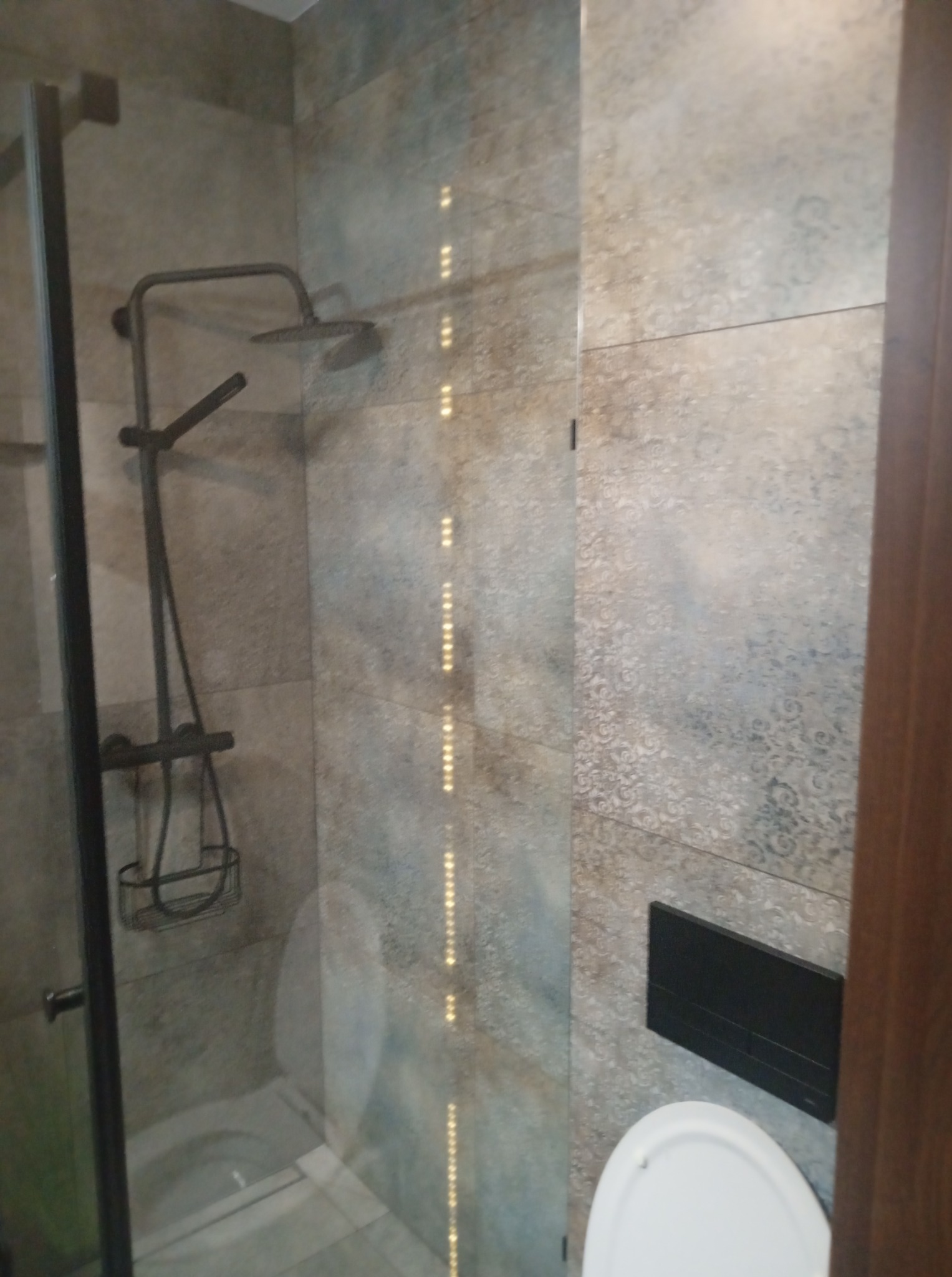 Nowoczesna łazienka z kabiną prysznicową typu walk-in, deszczownicą w kolorze grafitowym i toaletą. Płytki w odcieniach szarości z subtelnym wzorem.