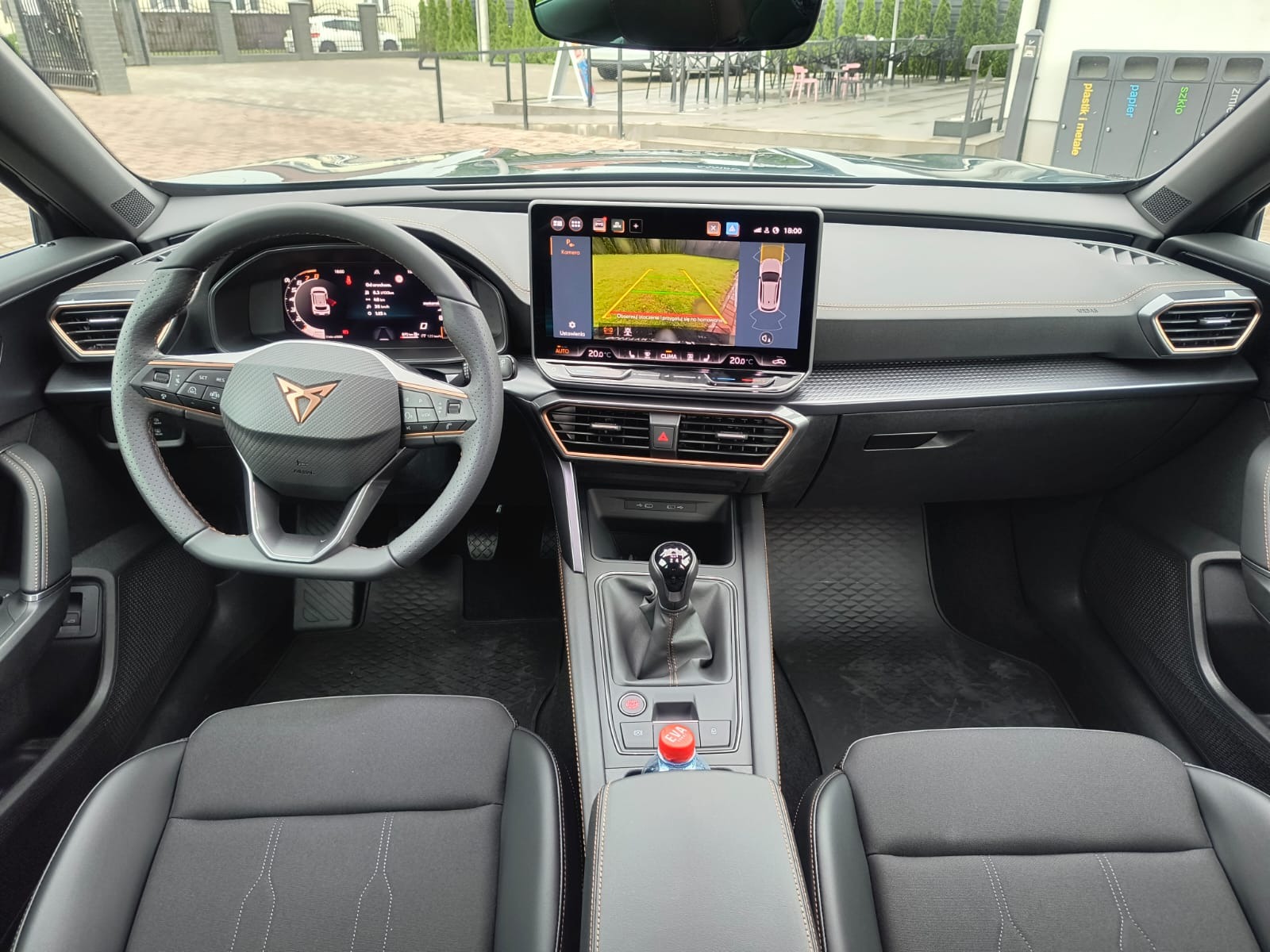 Wnętrze samochodu Cupra z widocznym systemem parkowania na ekranie, czarnymi fotelami i kierownicą z logo. W tle pojemniki na segregację odpadów.