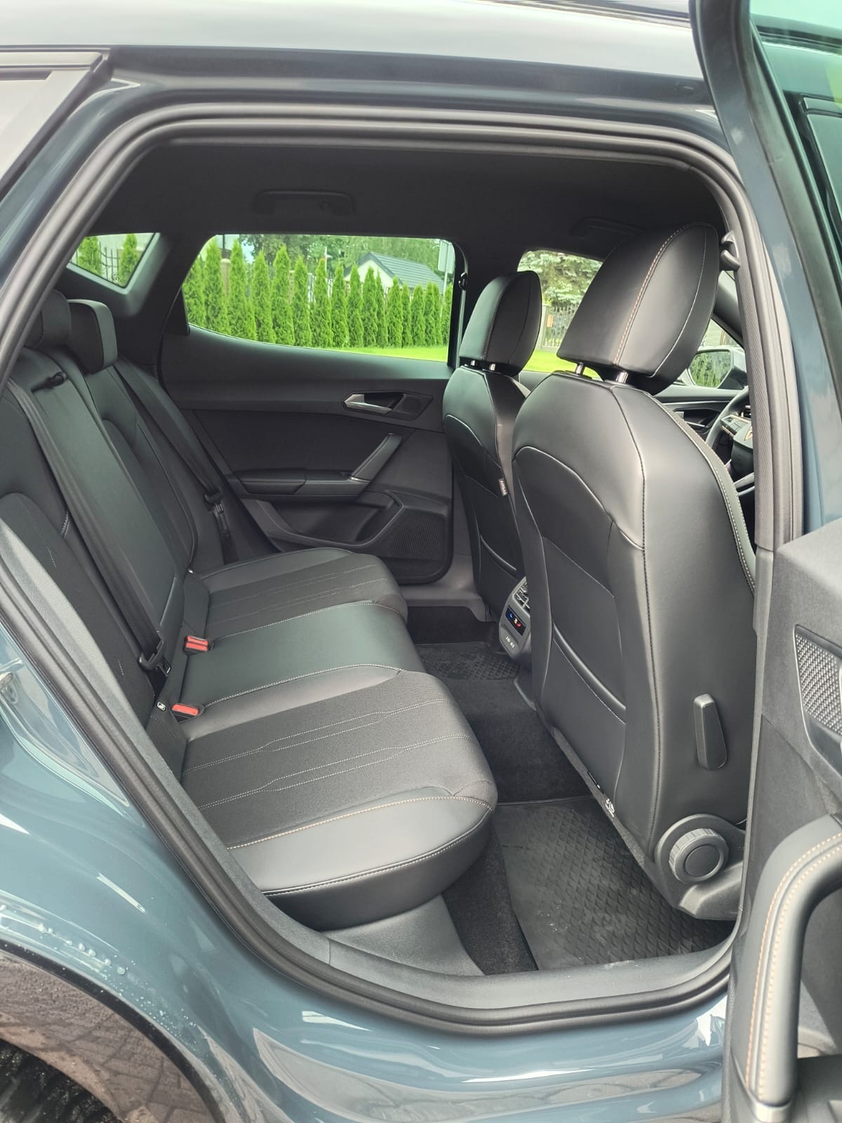 Wnętrze samochodu z otwartymi drzwiami, widok na tylną kanapę z czarnej skóry, zagłówki i dywaniki. Za oknem zieleń i budynek. Auto w kolorze szaro-niebieskim.