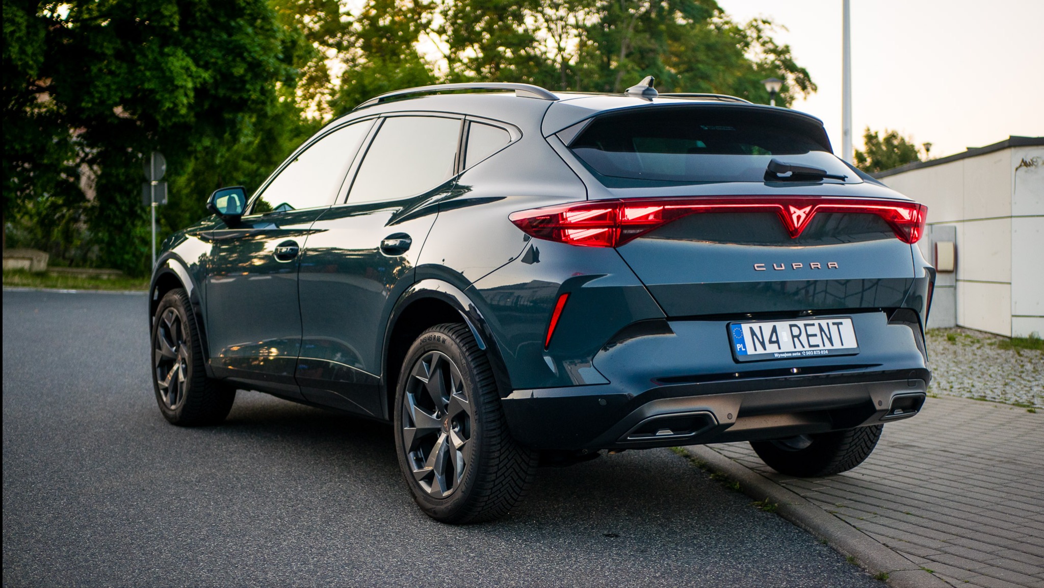 Ciemnozielony Cupra Formentor z tablicą rejestracyjną 'N4 RENT' stoi na ulicy. Widok z tyłu podkreśla nowoczesny design i światła LED. Tło stanowią drzewa i budynek.
