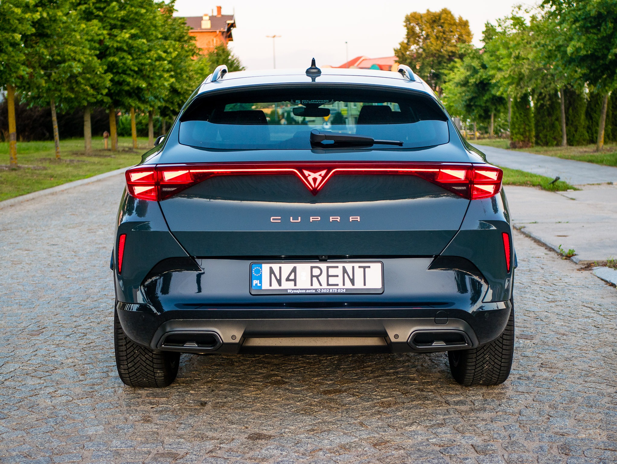Ciemny, lśniący Cupra Formentor z tablicą rejestracyjną 'N4 RENT' na tle brukowanej drogi i zieleni. Widok z tyłu, podkreślający nowoczesny design.