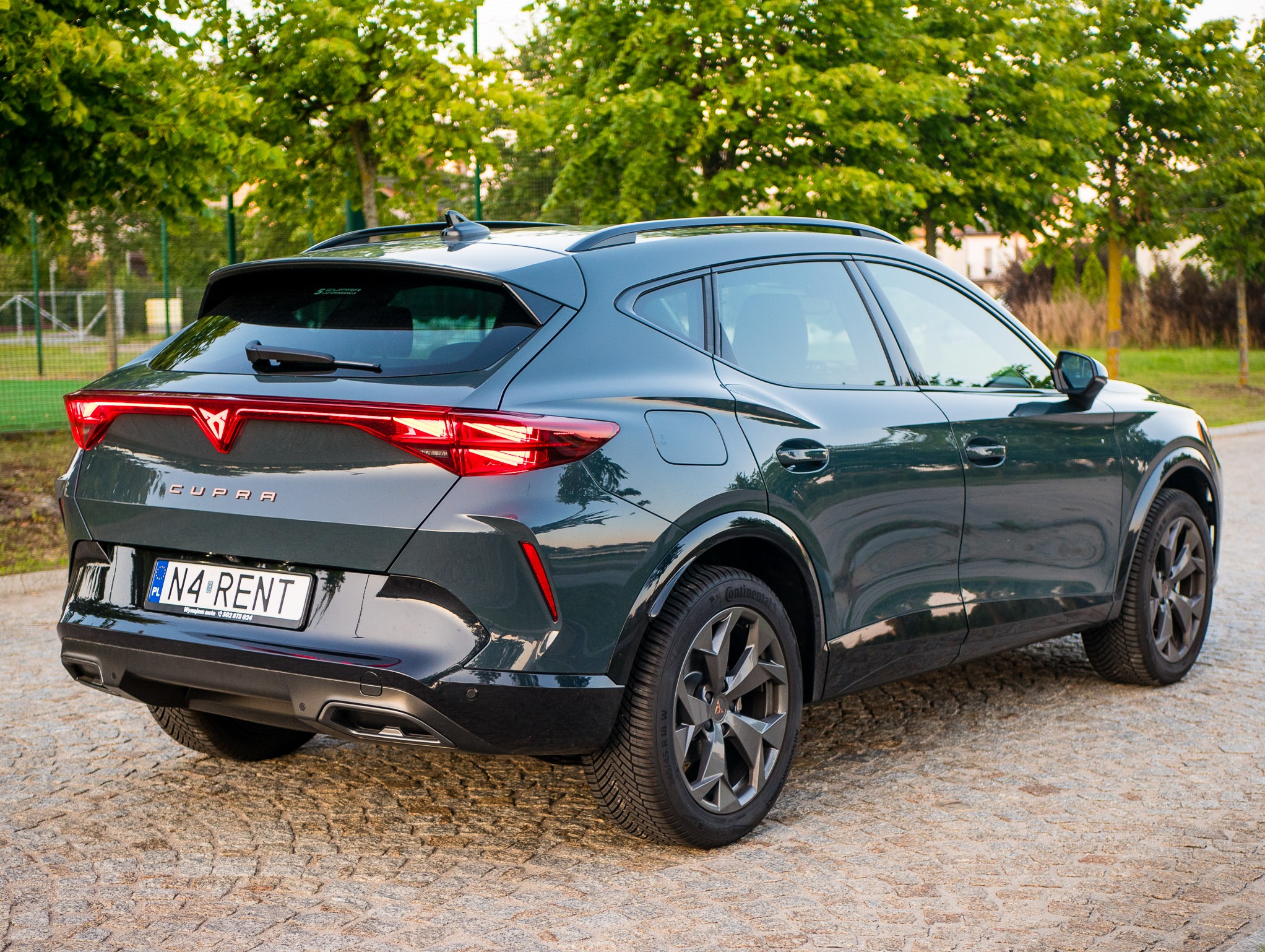 Zielony samochód Cupra Formentor z tablicą rejestracyjną 'N4 RENT' na tle zieleni. Widok z tyłu, oświetlenie naturalne, kostka brukowa.