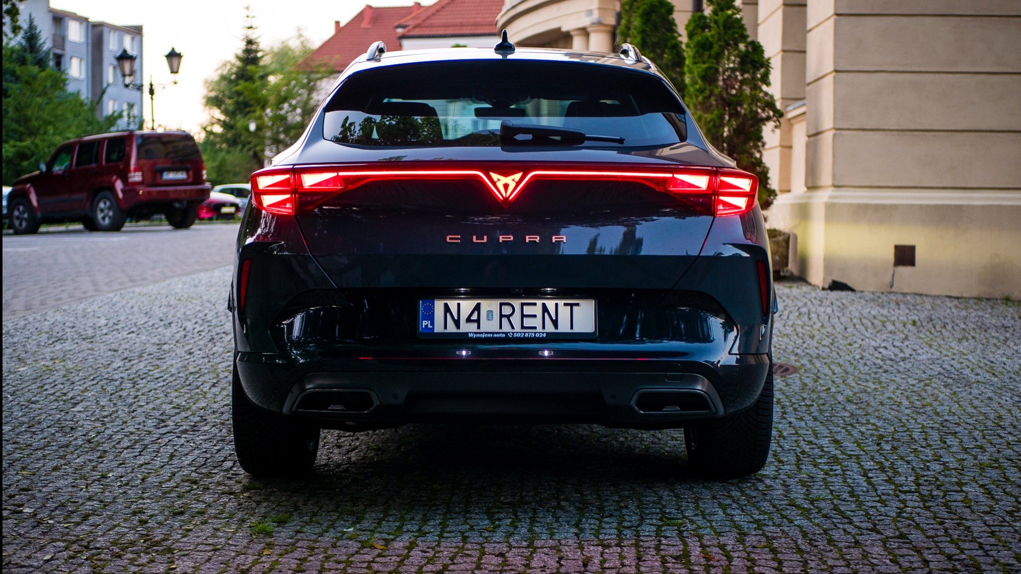Ciemny samochód Cupra z włączonymi światłami pozycyjnymi, widok z tyłu, z tablicą rejestracyjną 'N4 RENT' na brukowanym podłożu.