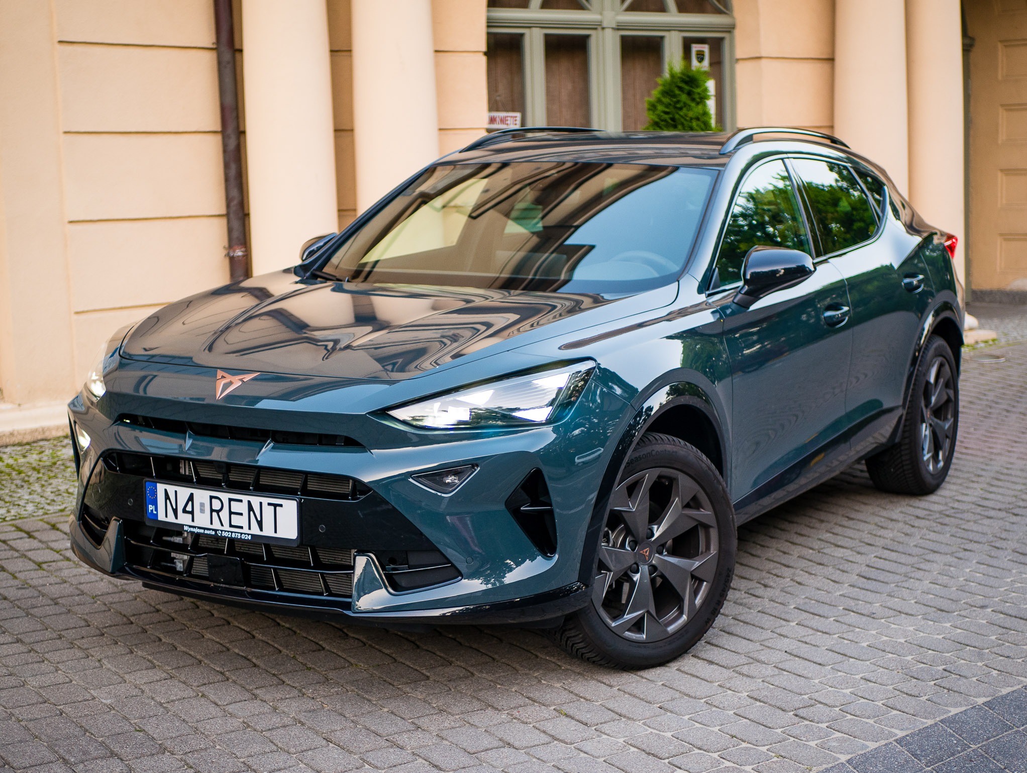 Zielony samochód Cupra Formentor z tablicą rejestracyjną 'N4 RENT' na brukowanej ulicy, zaparkowany przed budynkiem. Widok z przodu, lekko z dołu.