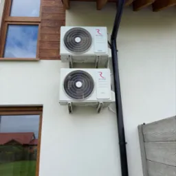 Dwa białe klimatyzatory Rotenso na elewacji budynku, zamontowane jeden nad drugim, obok rynny. Widoczne okna z drewnianą ramą i fragment drewnianego podbitki.