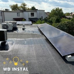 WD INSTAL - Instalacja paneli słonecznych na płaskim dachu budynku, widoczne również jednostki klimatyzacyjne i zabudowa miejska w tle, pochmurne niebo.