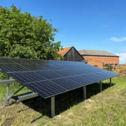 Instalacja fotowoltaiczna na łące, w tle budynek gospodarczy z cegły i sterta siana pod błękitnym niebem. Panele ustawione pod kątem na metalowej konstrukcji.