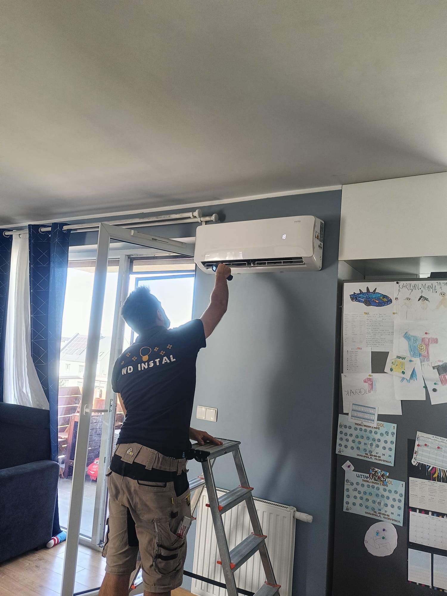 Instalator na drabinie montuje klimatyzator Rotenso na ścianie w pokoju z lodówką ozdobioną rysunkami dziecięcymi i kalendarzem.