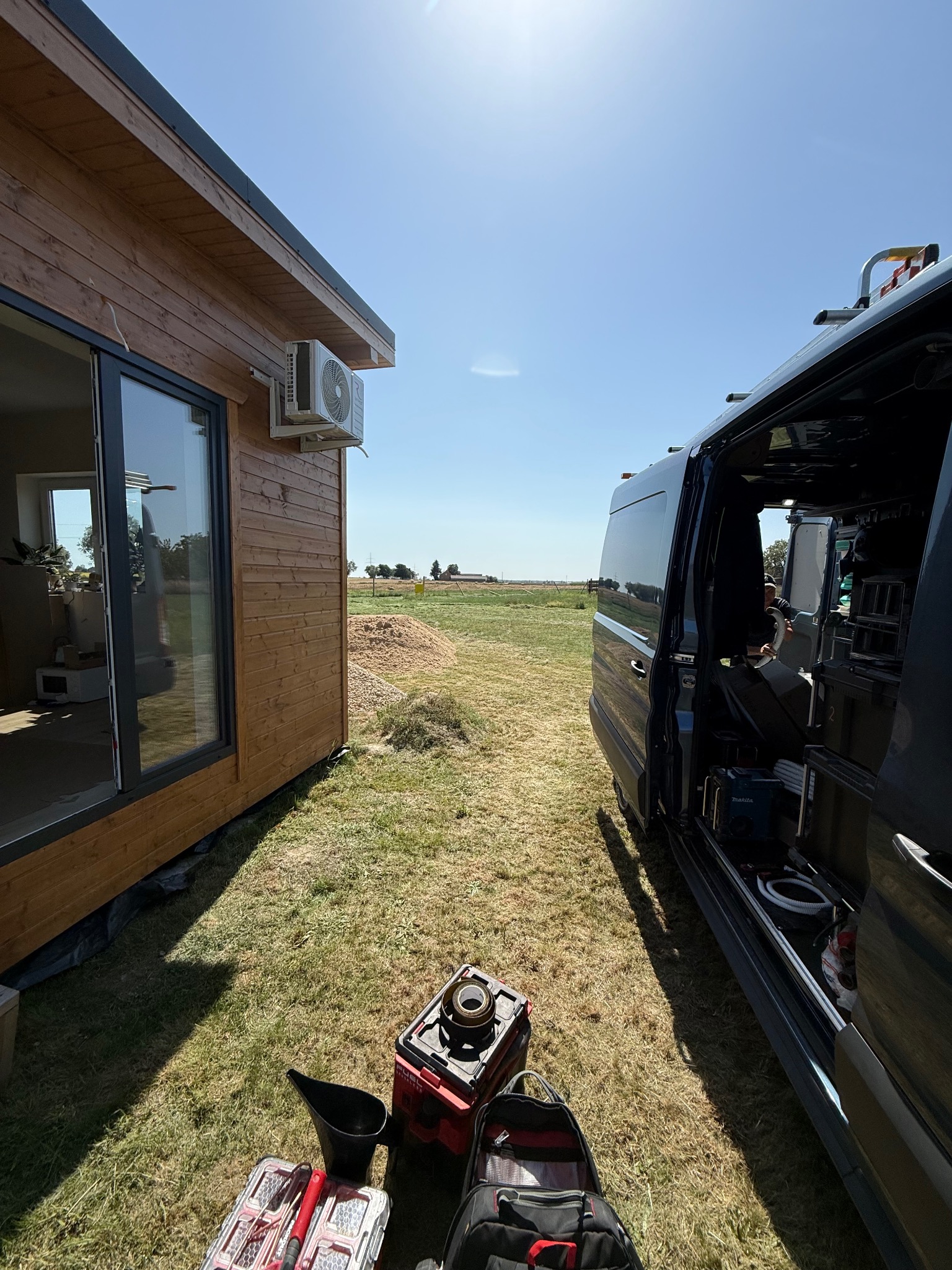 Klimatyzator Rotenso zamontowany na drewnianym domku, widok z poziomu trawy, z narzędziami i otwartym busem serwisowym w słoneczny dzień.