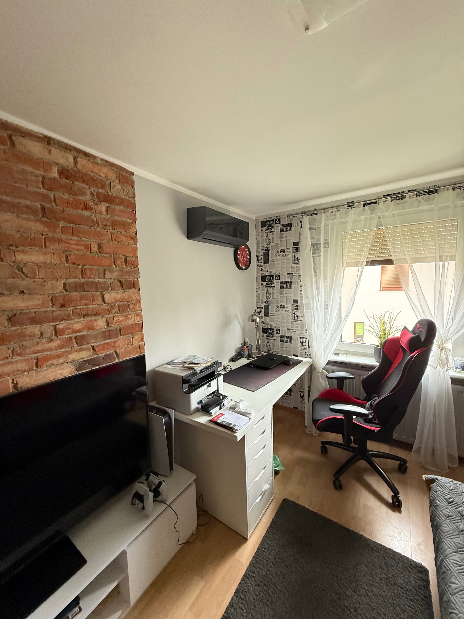Szara klimatyzacja Rotenso na ścianie w pokoju z ceglaną ścianą, biurkiem, fotelem gamingowym i tapetą w gazety. Widok na okno z białymi firanami.