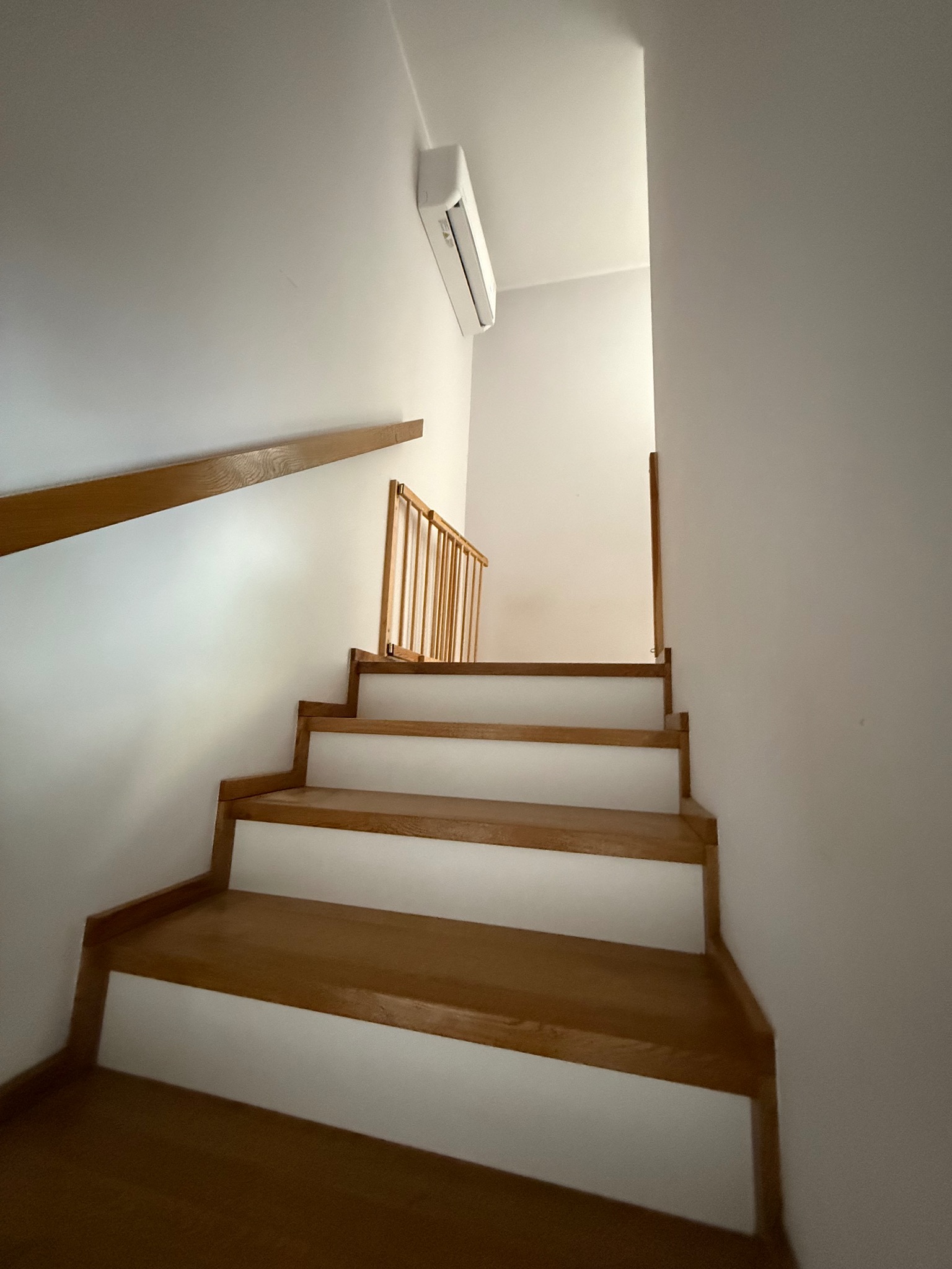 Biały klimatyzator ścienny Rotenso nad drewnianymi schodami z barierką w jasnym wnętrzu. Ujęcie z dołu, podkreślające wysokość pomieszczenia.