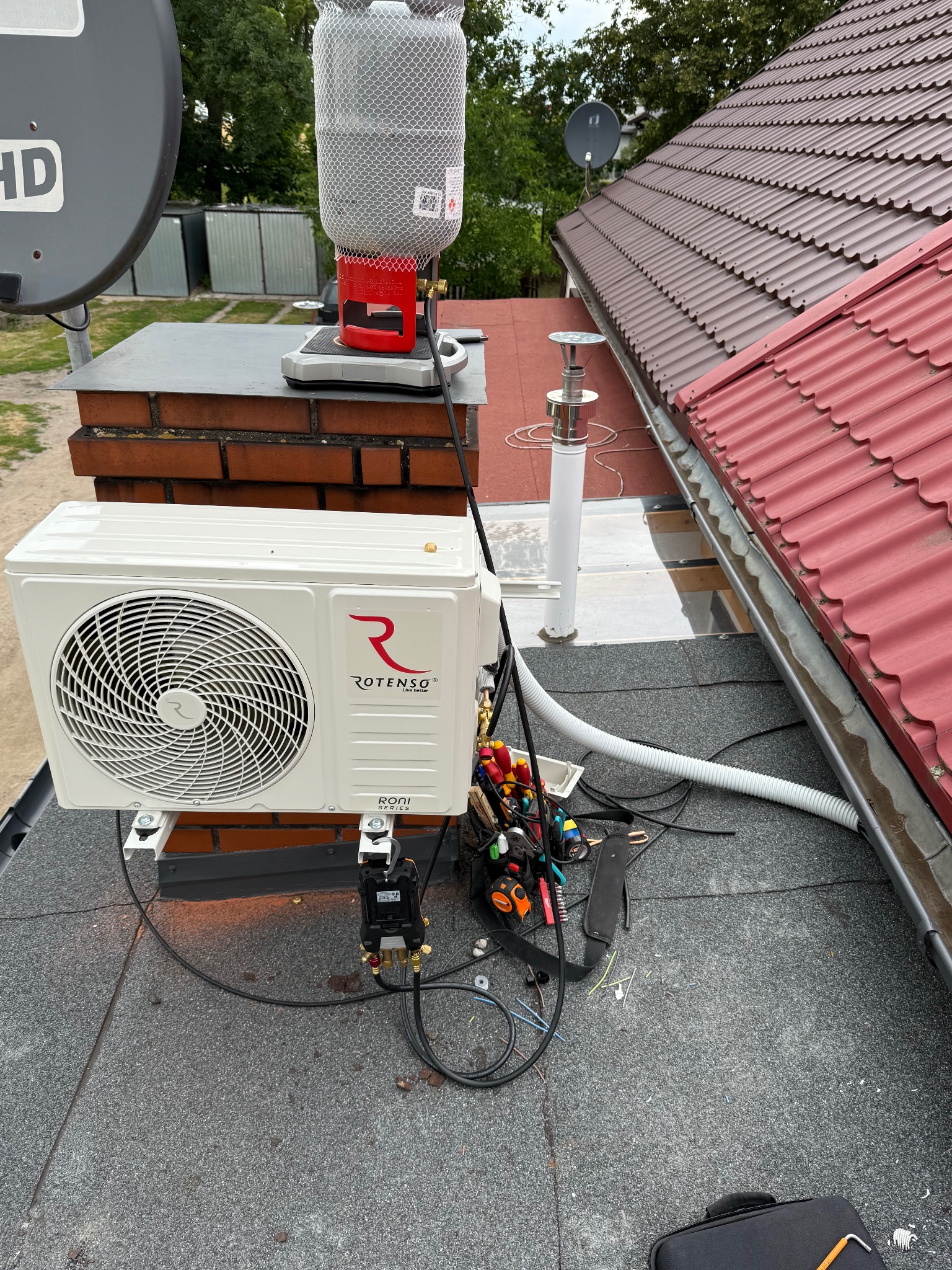 Biała jednostka zewnętrzna klimatyzacji Rotenso zamontowana na dachu, obok narzędzia i butla z gazem. Widok na dach z czerwoną dachówką i anteny satelitarne.