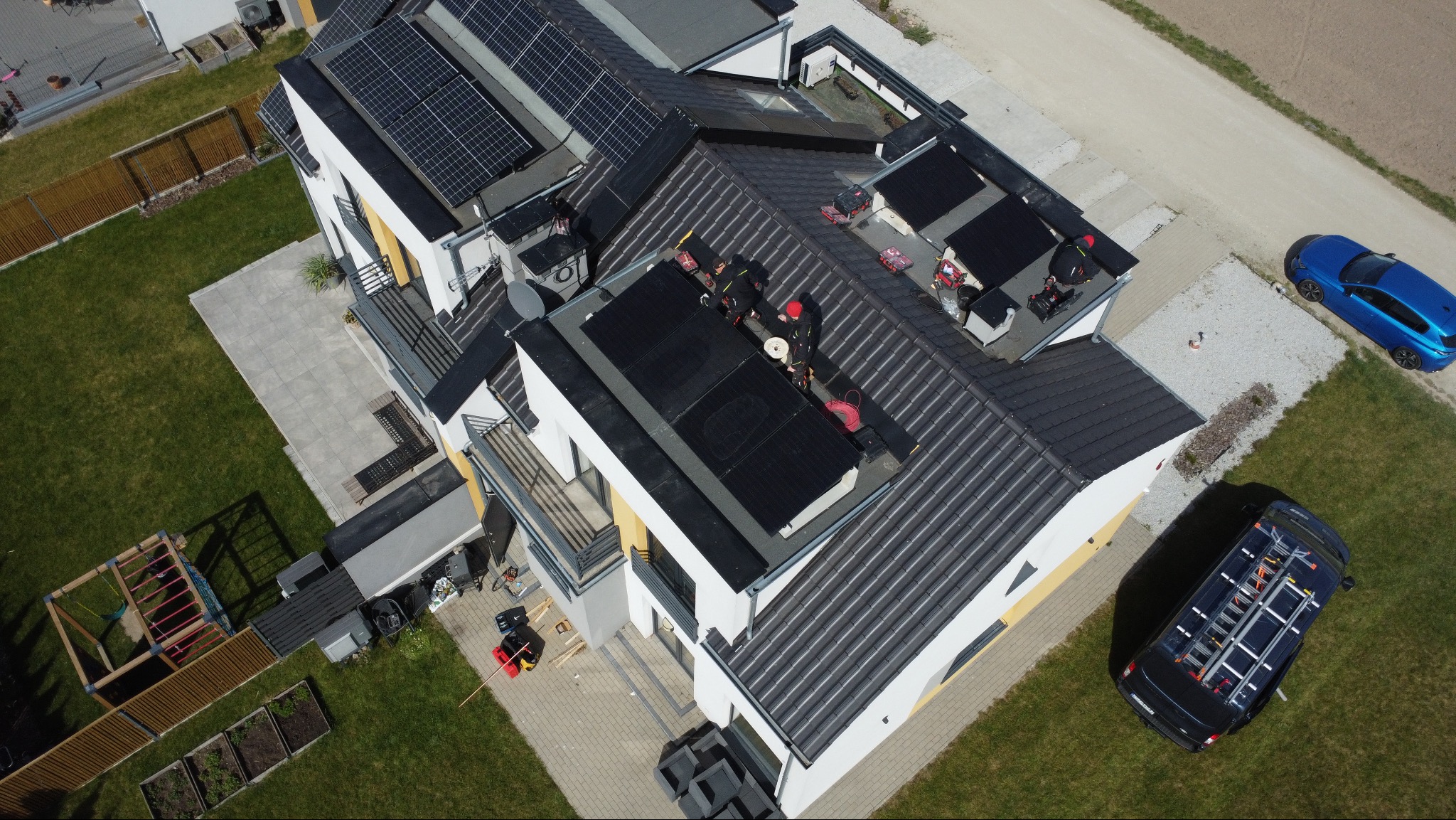 Montaż paneli słonecznych na dachu nowoczesnego domu z bliska, widok z drona. Ekipa w czarnych strojach instaluje panele, obok zaparkowany samochód z drabiną.