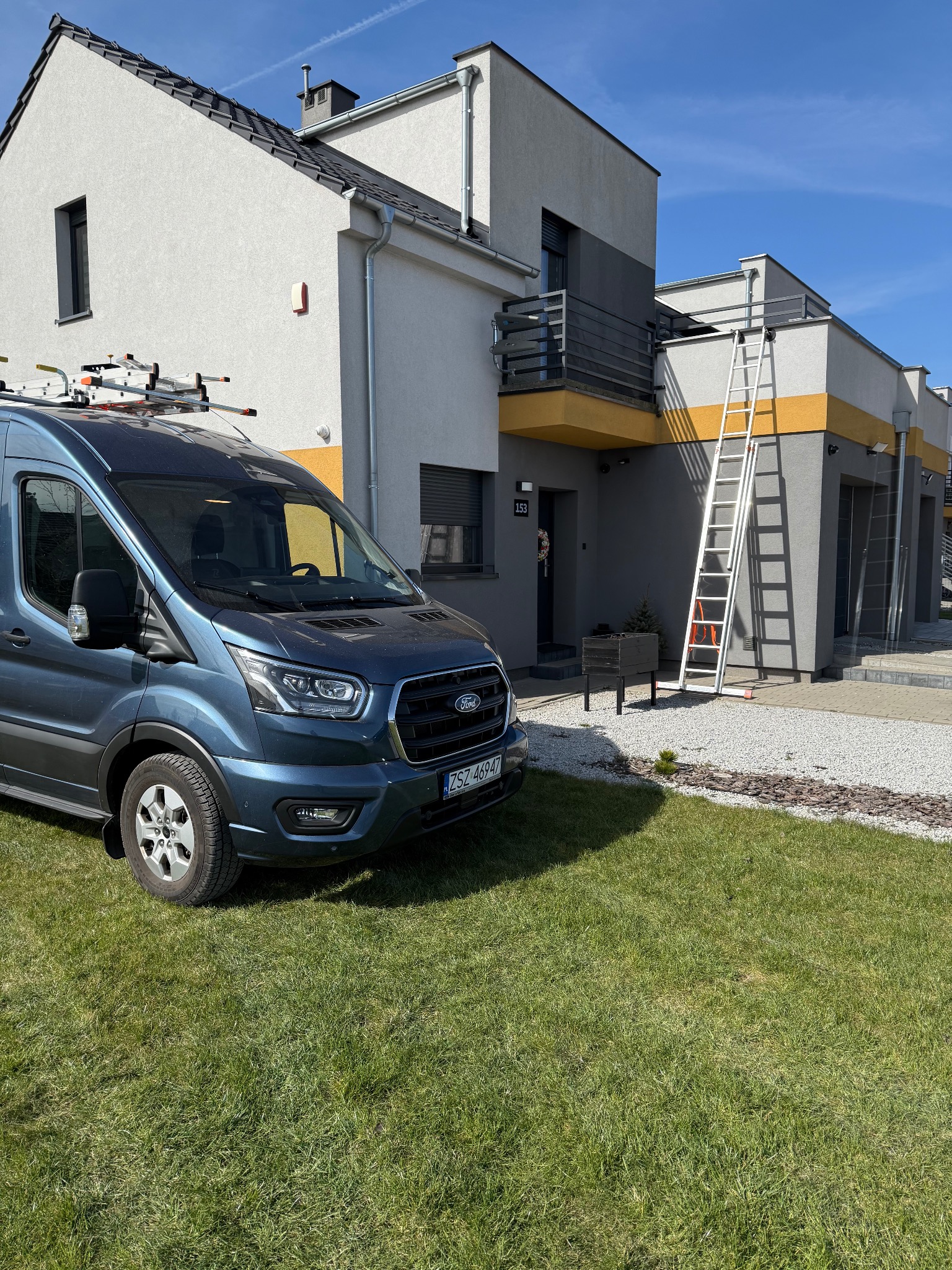 Niebieski bus Ford Transit z drabiną na dachu stoi na trawniku przed nowoczesnym domem z elementami w kolorze żółtym. Drabina oparta o ścianę budynku.