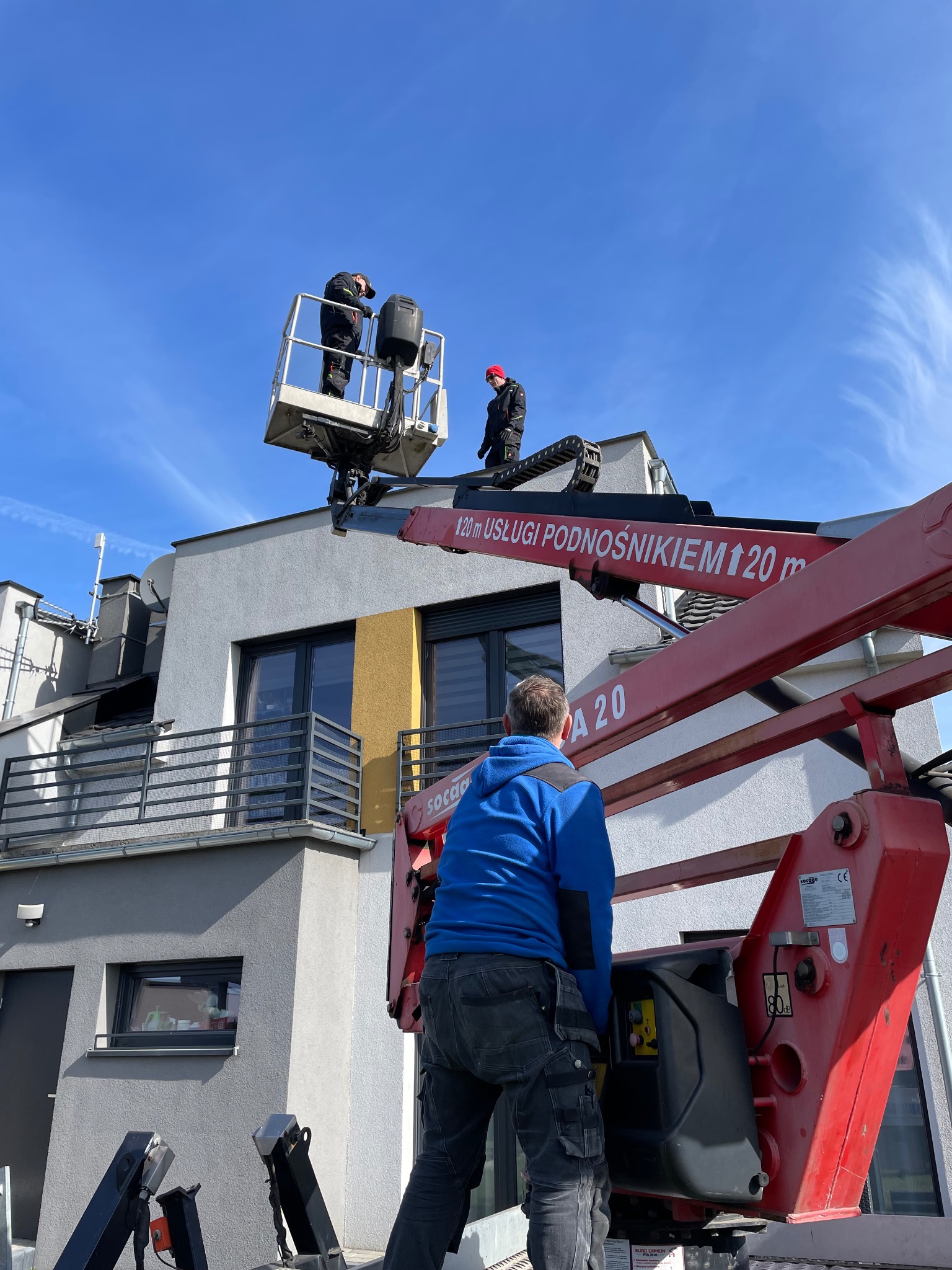 Praca na wysokościach: dwaj monterzy w koszu podnośnika na dachu budynku, nadzorowani przez osobę z dołu. Czerwone ramię podnośnika z napisem 'USŁUGI PODNOŚNIKIEM 20 m'.
