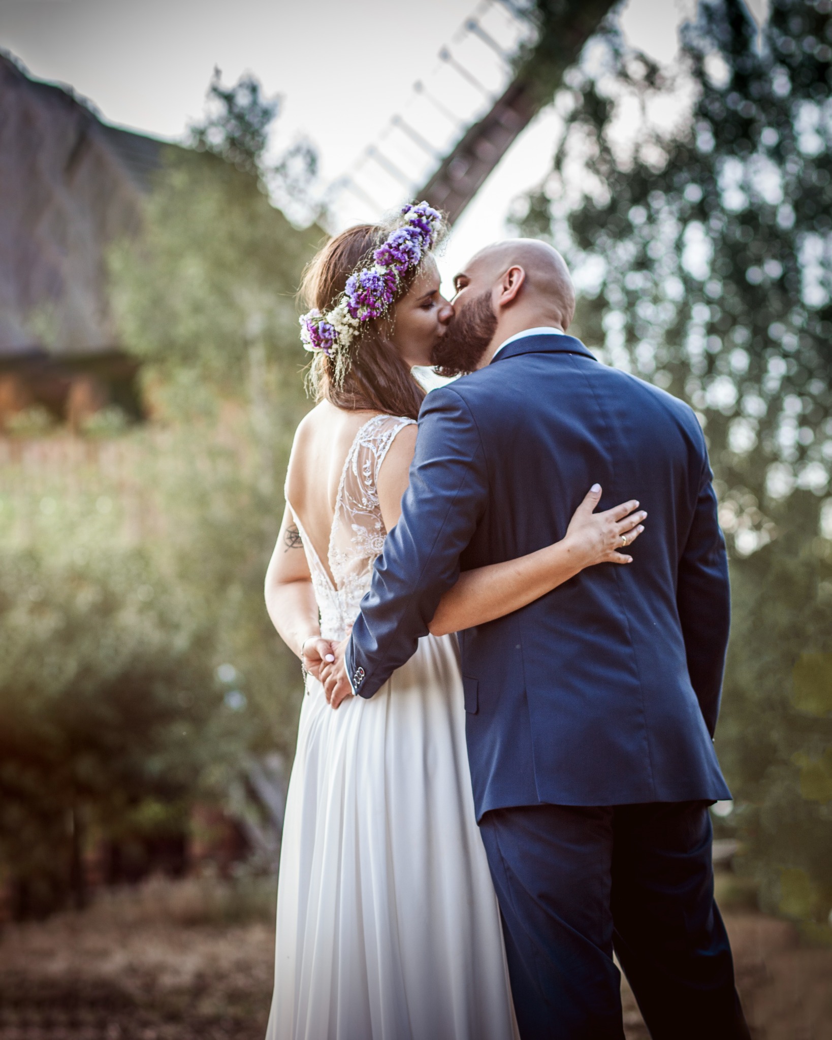 Pocałunek pary młodej na tle wiatraka. Panna młoda w sukni z odkrytymi plecami i wiankiem z fioletowych kwiatów, pan młody w granatowym garniturze.