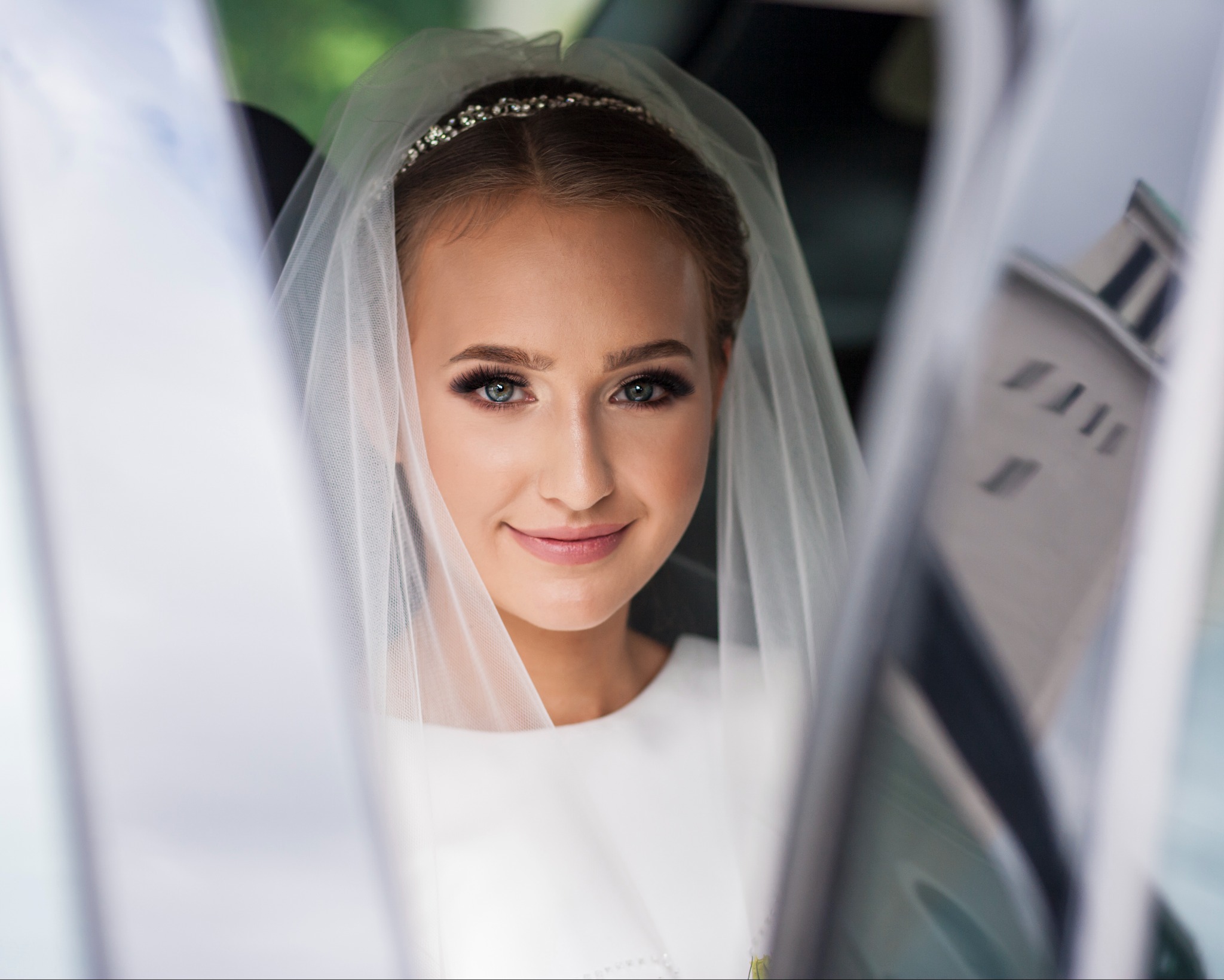 Uśmiechnięta panna młoda w welonie i diademie, portret z bliska w samochodzie, z widocznym fragmentem budynku w tle przez okno.