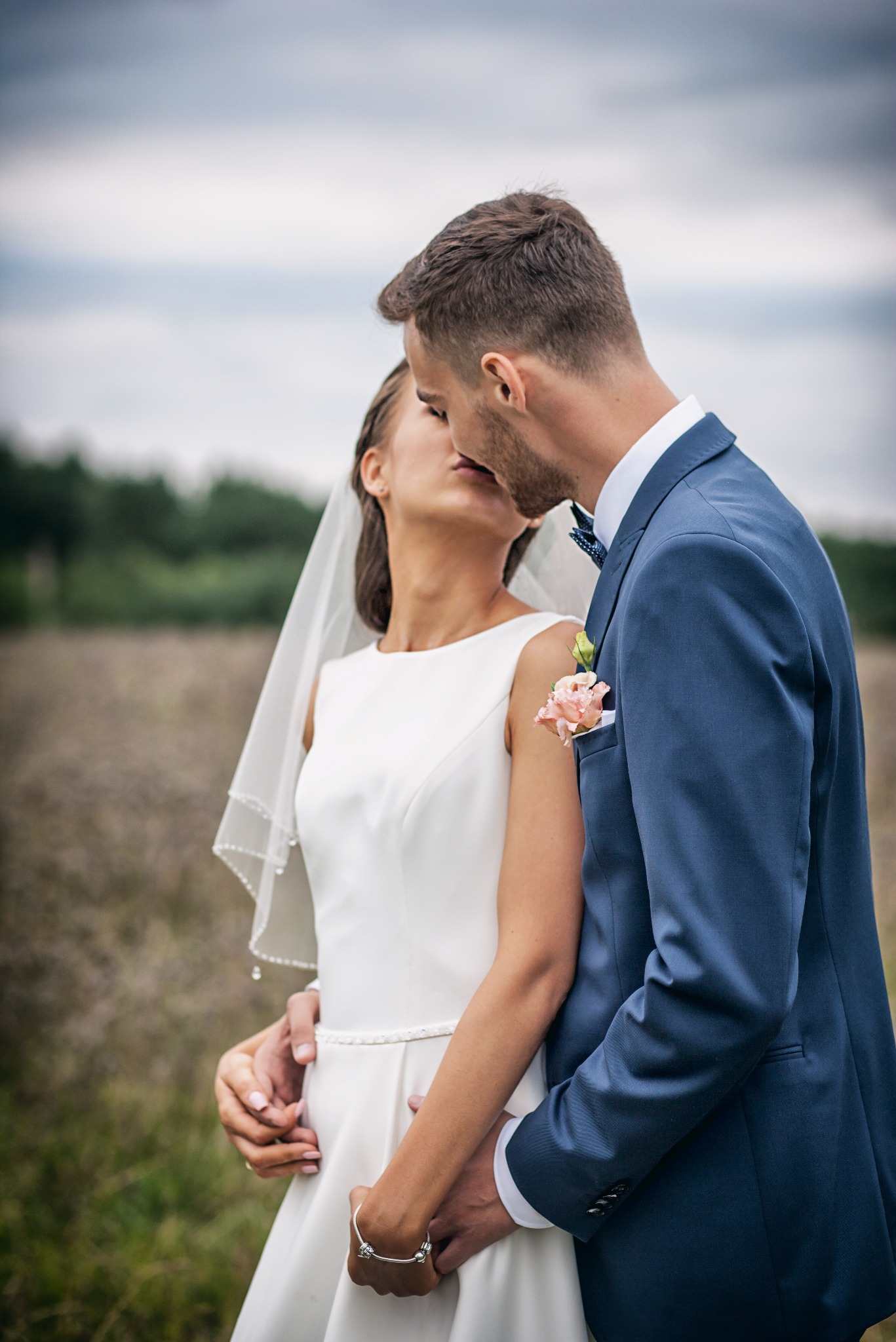 Młoda para w objęciach, delikatny pocałunek na tle łąki. Panna młoda w prostej sukni z welonem, pan młody w granatowym garniturze z butonierką.