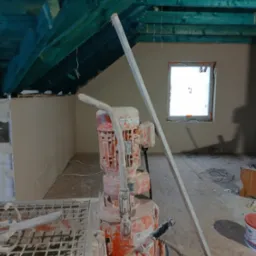 Wnętrze w trakcie prac tynkarskich. Widoczna maszyna do tynkowania, ściany pokryte świeżym tynkiem oraz drewniana konstrukcja dachu. Dwa okna w tle.