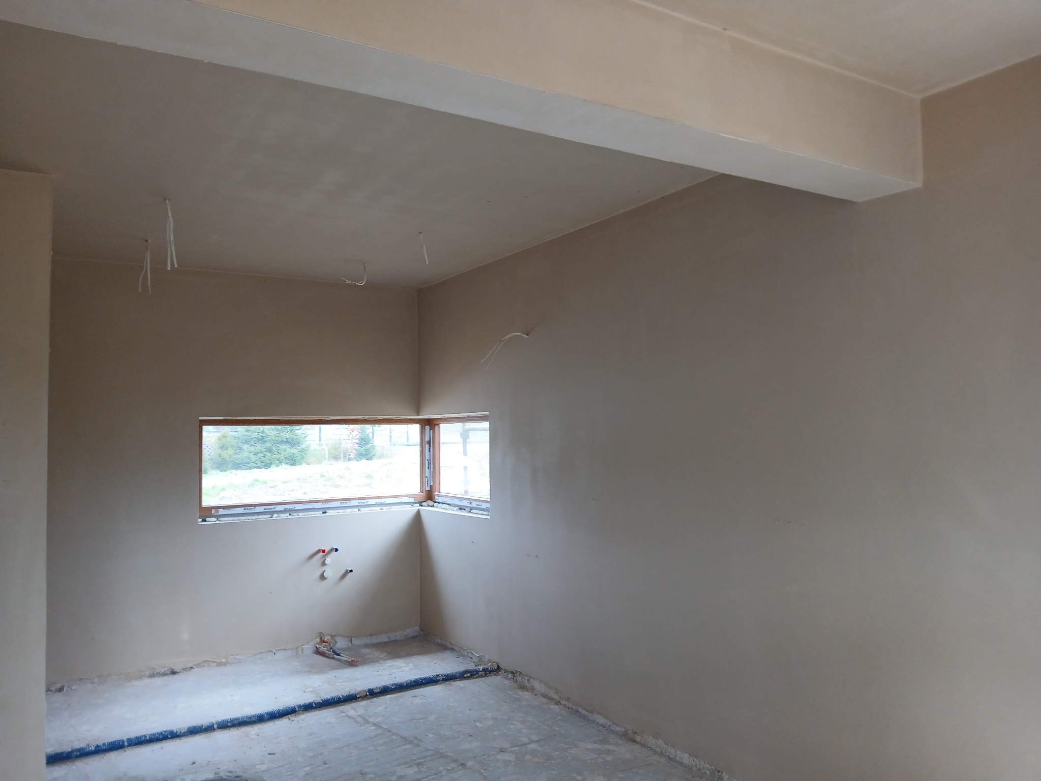 Wnętrze pokoju po nałożeniu tynków. Widoczne gładkie ściany, okno narożne i wystające przewody instalacji. Podłoga przygotowana do dalszych prac wykończeniowych.