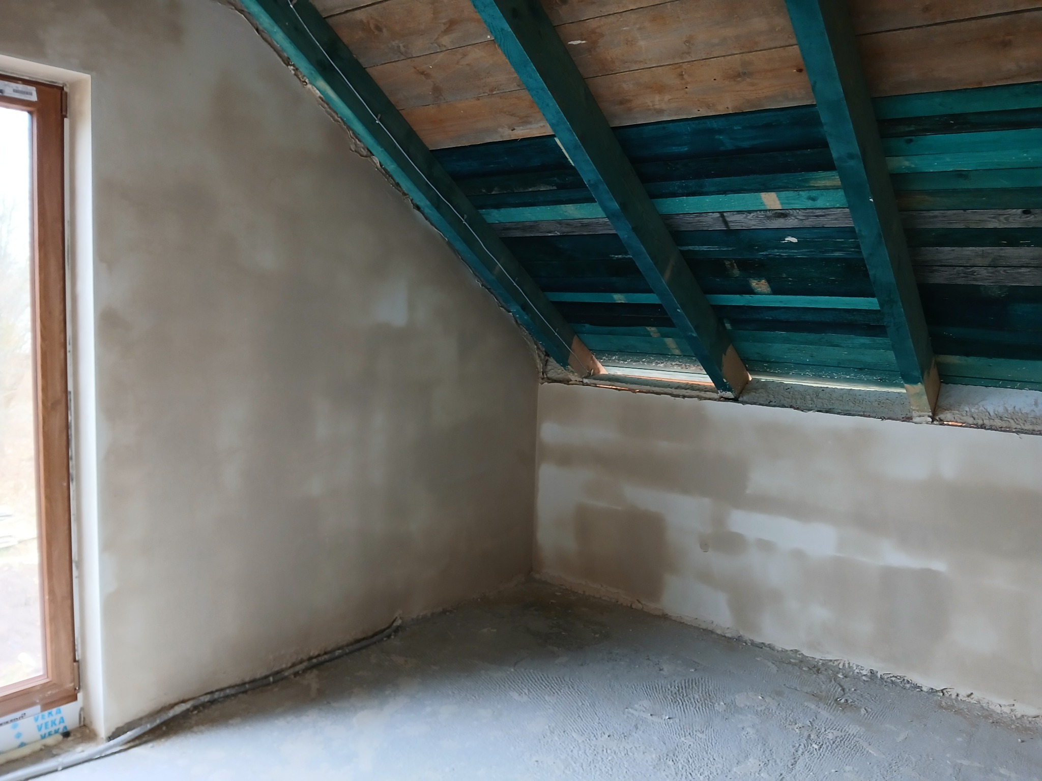 Wnętrze poddasza po nałożeniu tynków, widoczne ściany w jasnym kolorze, drewniana konstrukcja dachu pomalowana na turkusowo, okno z drewnianą ramą.